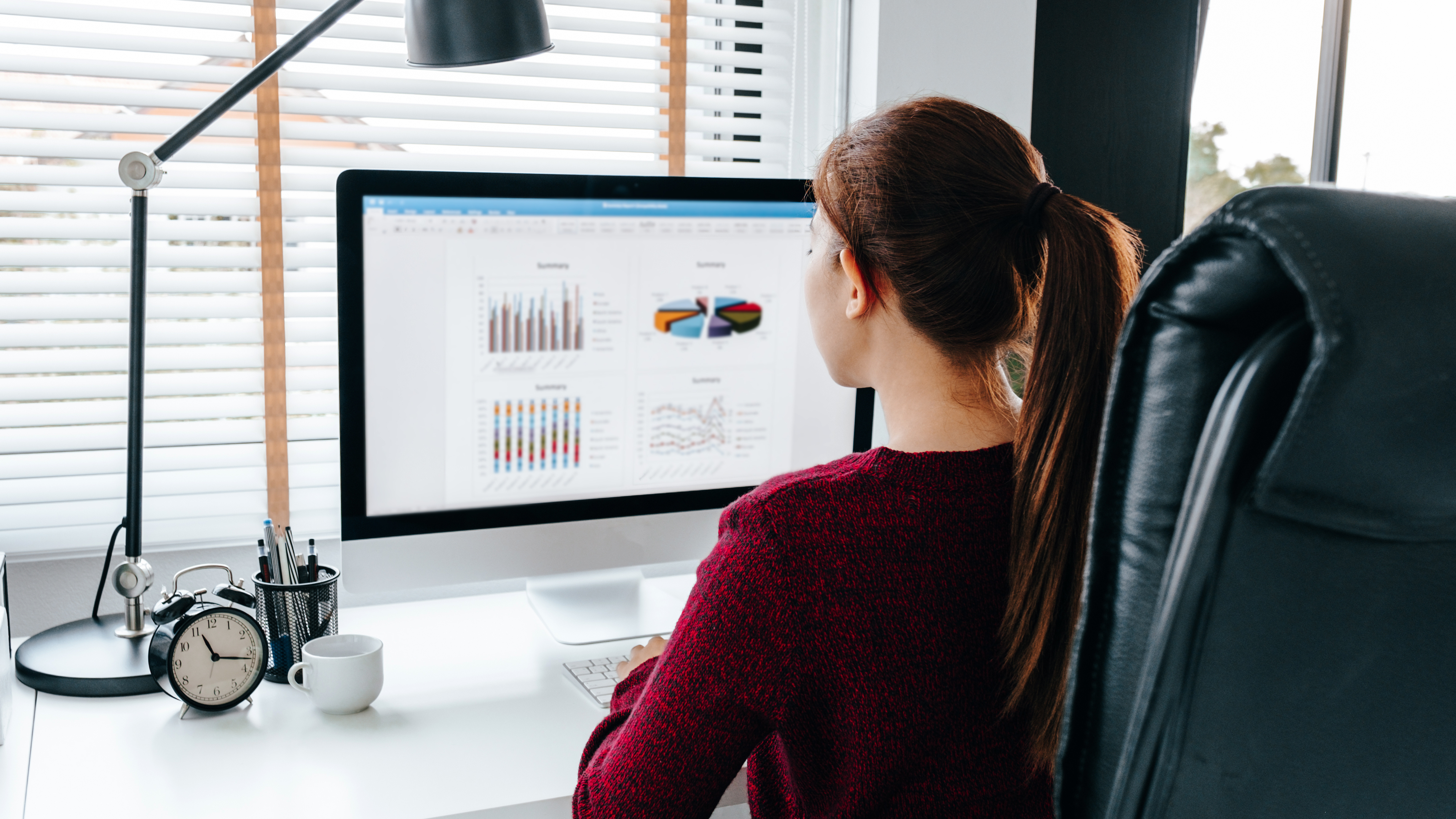 Woman working at a computer with graphs on screen, sitting at a desk with a lamp, clock, and plant.