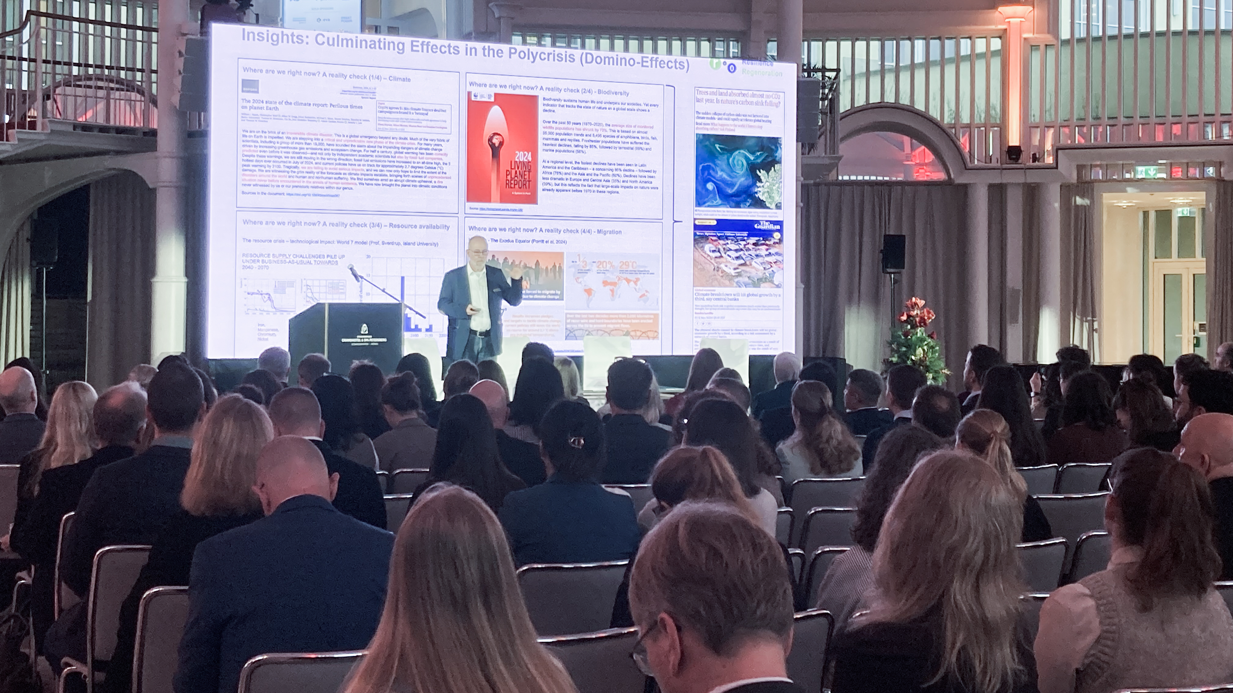 Conference speaker presents slide on polycrisis effects to seated audience in ornate, high-ceiling venue.