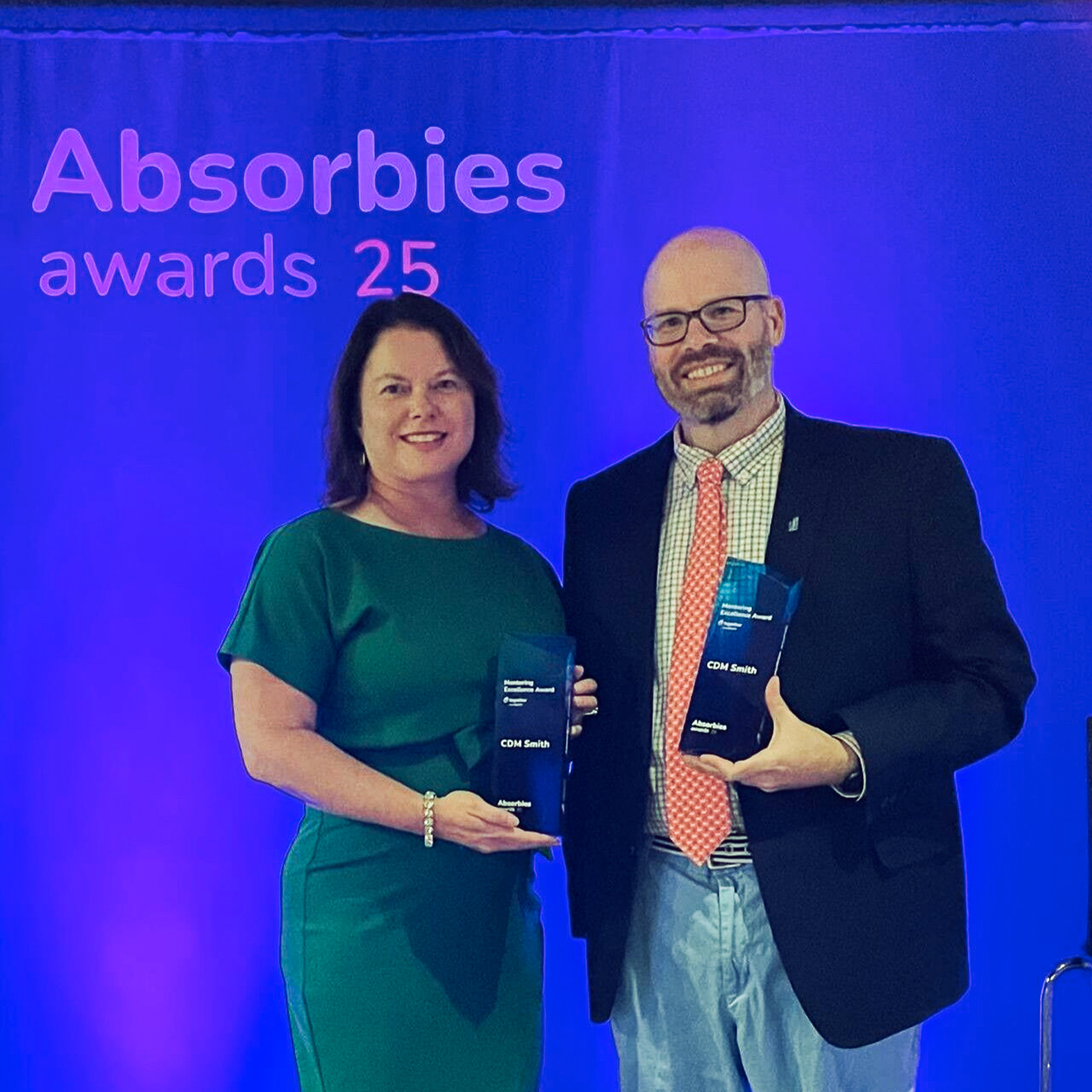 Two people holding awards at the Absorbies Awards 25 event, standing in front of a blue backdrop.