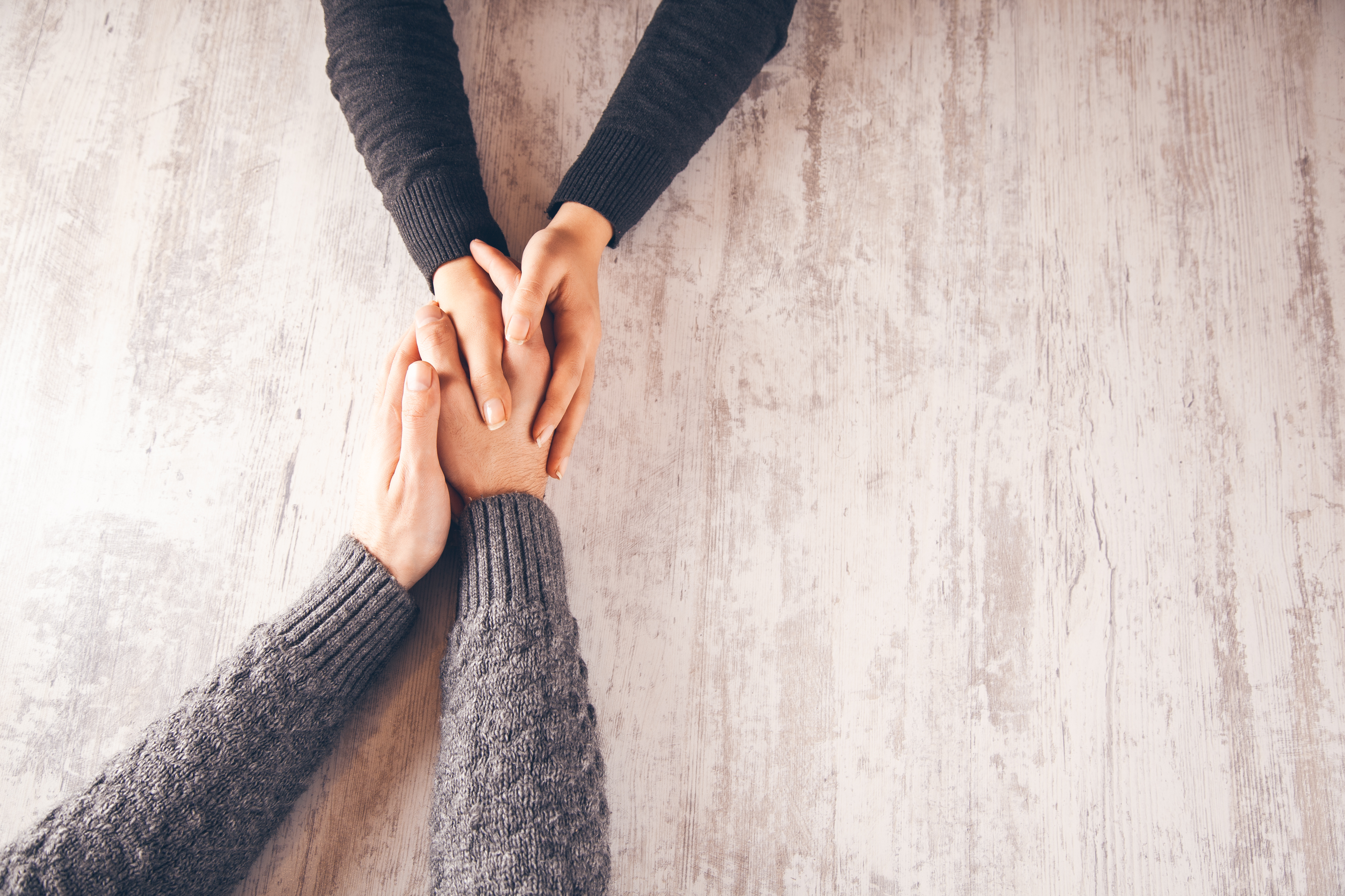 Two pairs of hands are clasped together on a wooden surface, expressing comfort and support.