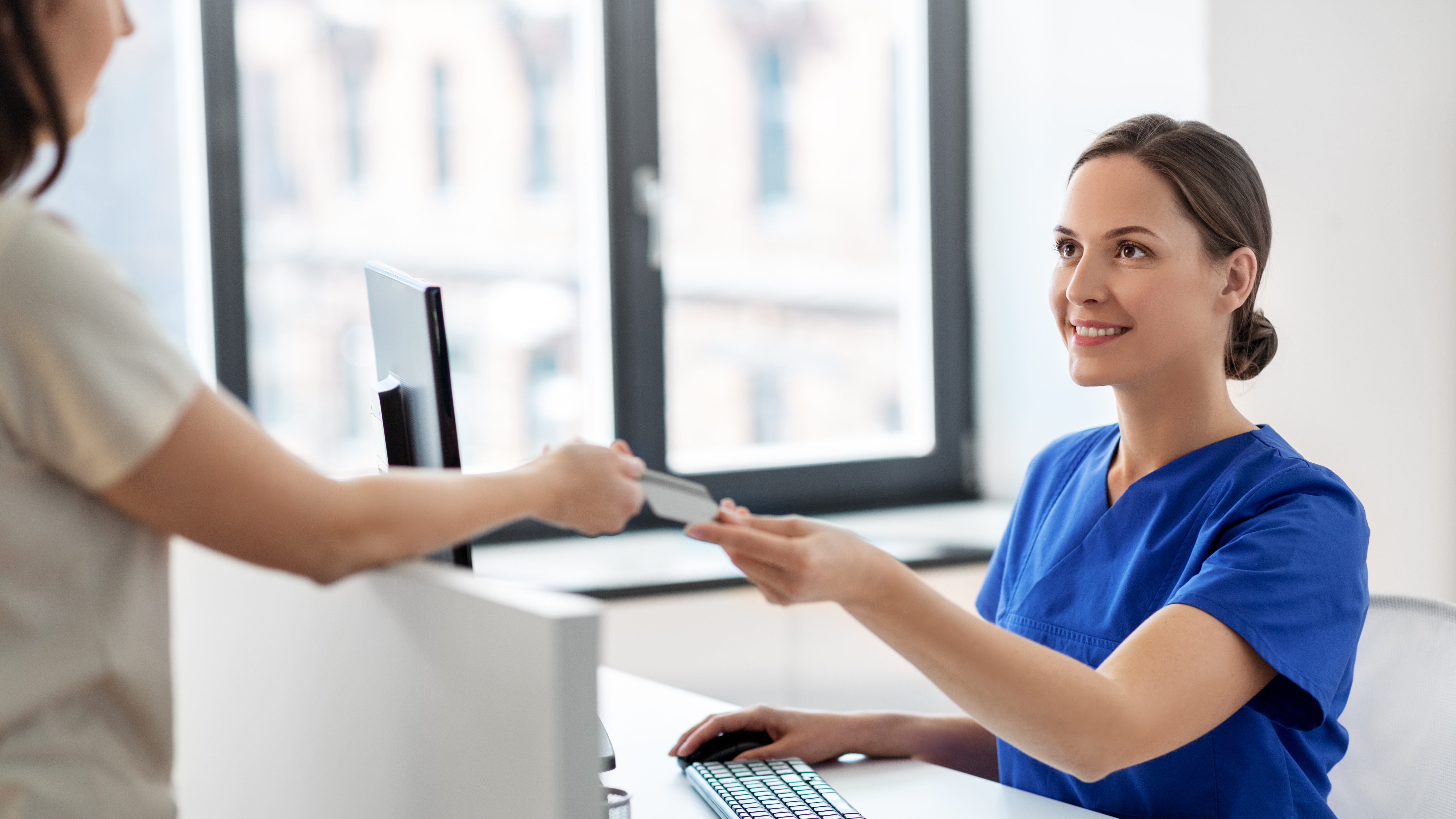 Healthcare worker in scrubs hands ID card to another person across a reception desk.