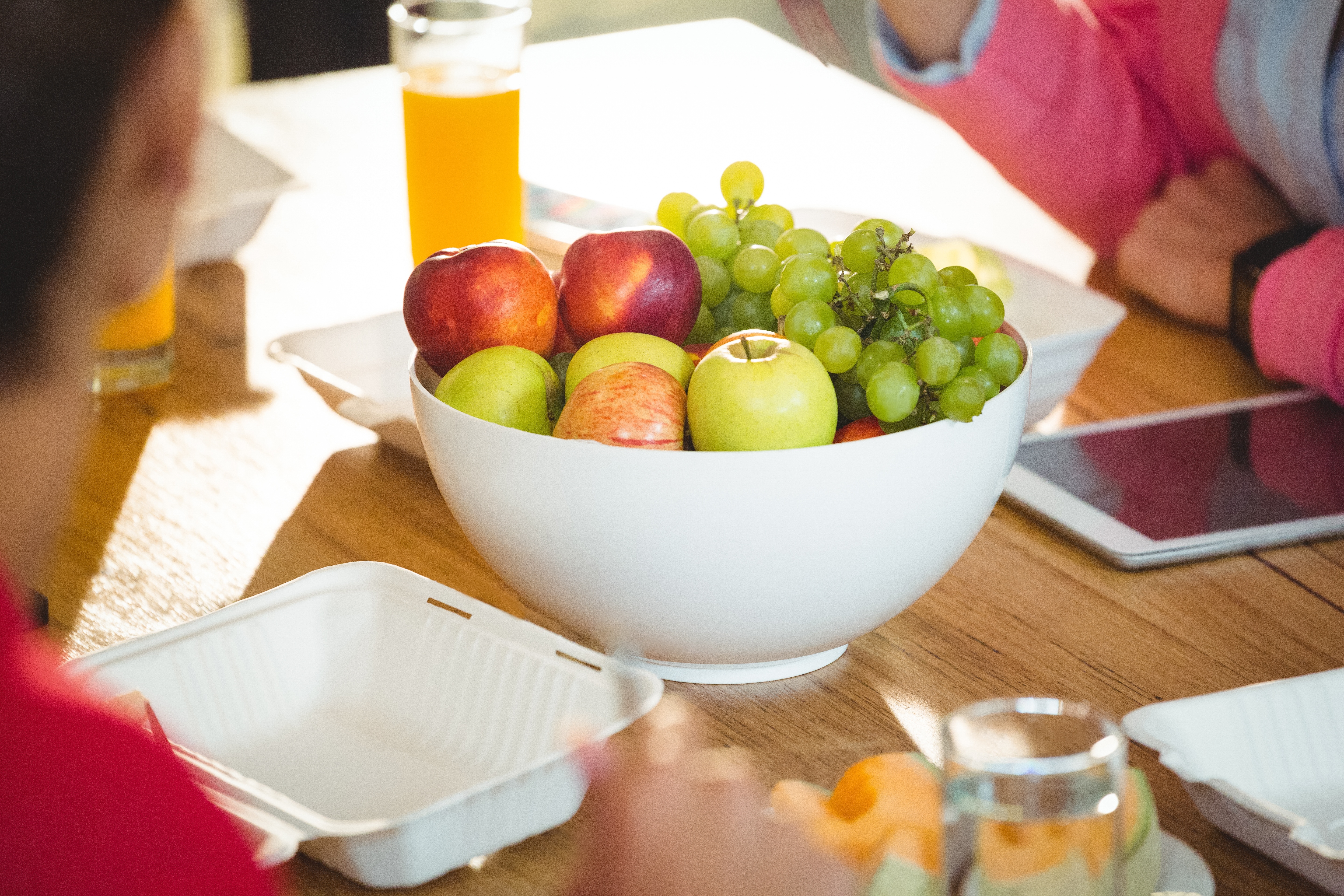 A white bowl of fresh fruit including apples and grapes on a wooden table surrounded by office items like glasses and trays.