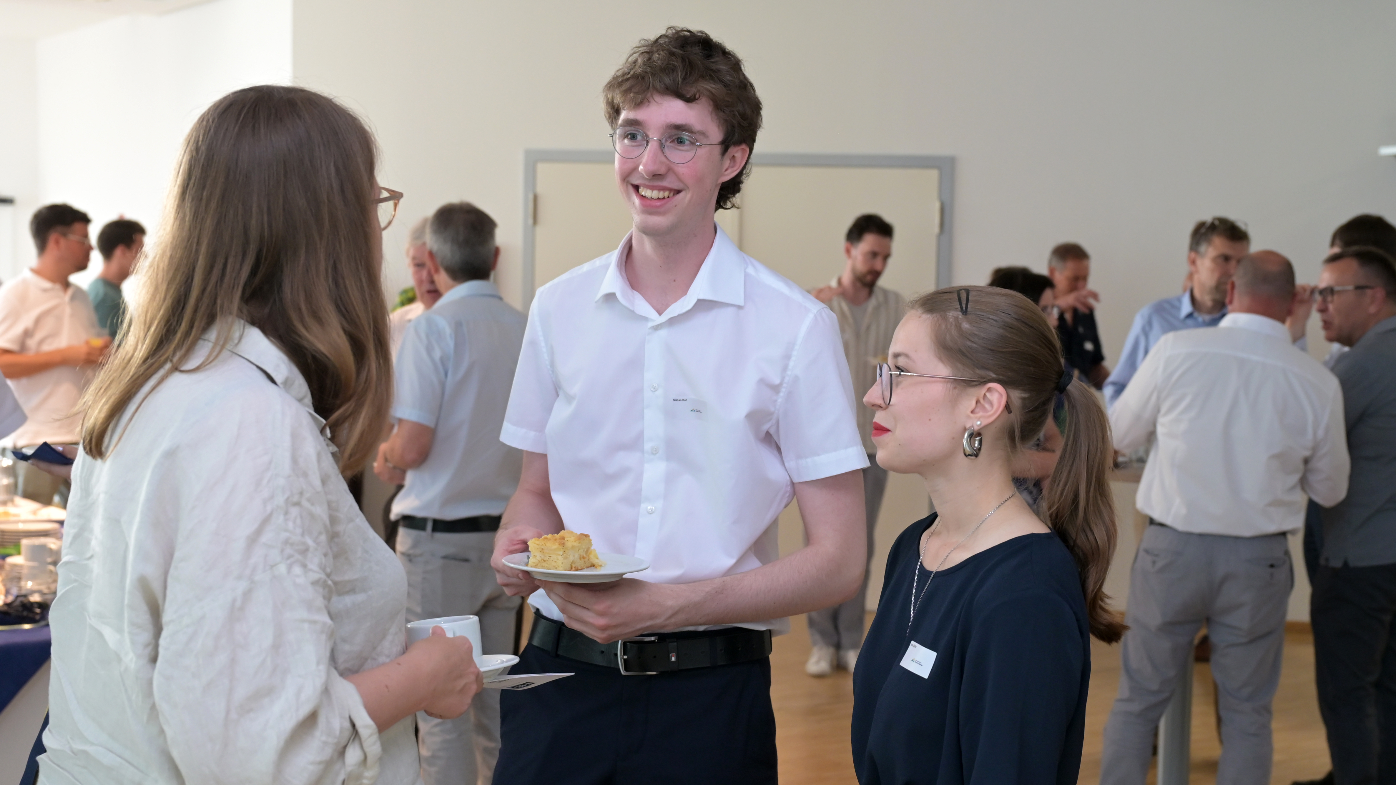 A group of people engaged in conversation during an indoor social event, with tables of refreshments visible in the background.