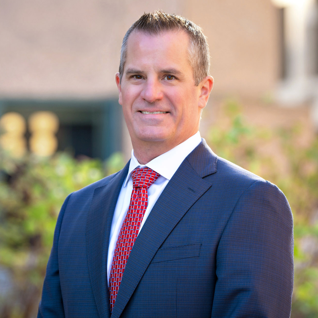 Person in a blue suit and red tie stands outdoors with blurred trees and buildings in the background.