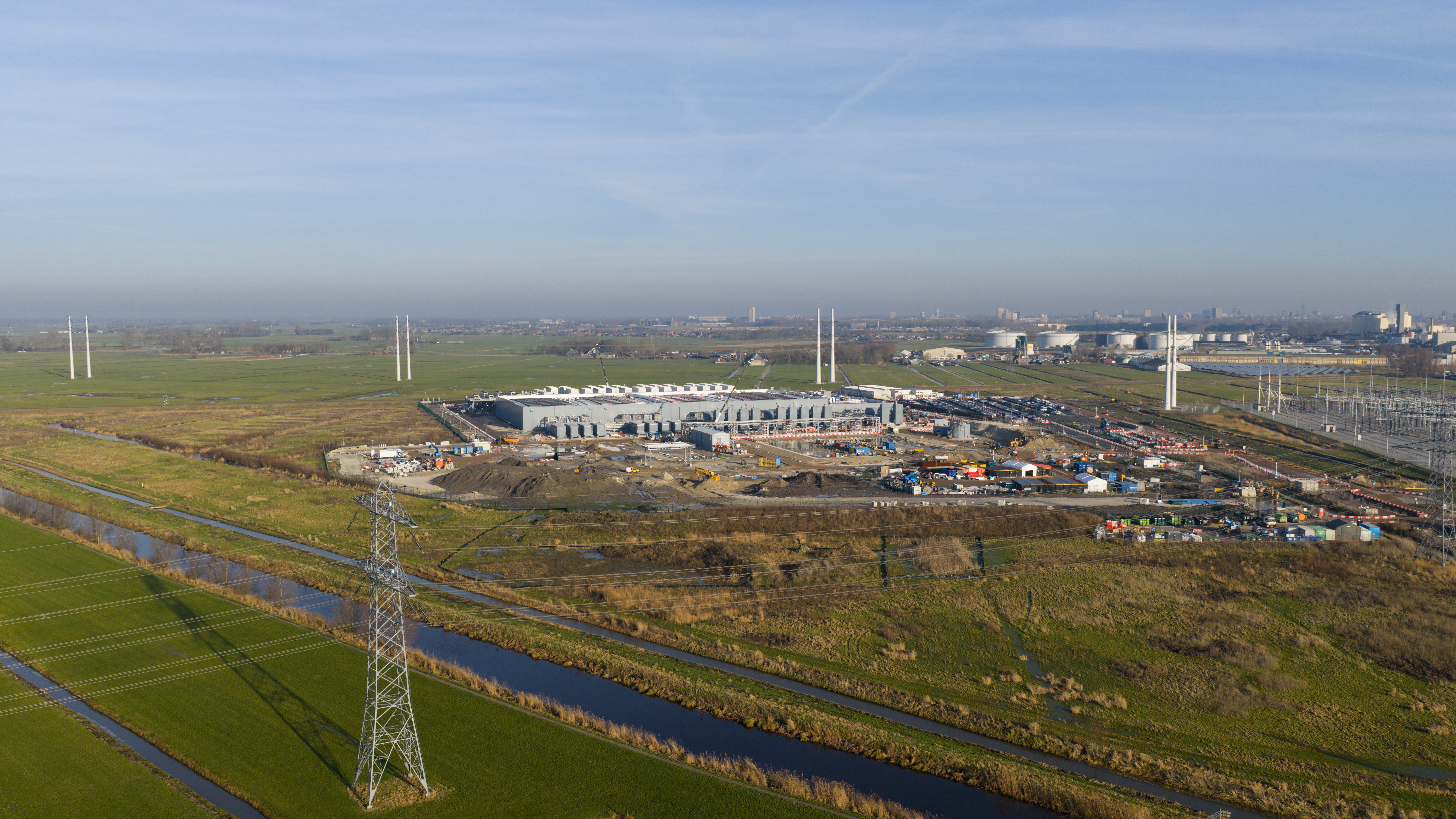 Aerial view of a data center under construction surrounded by green fields and power lines, with infrastructure development visible.