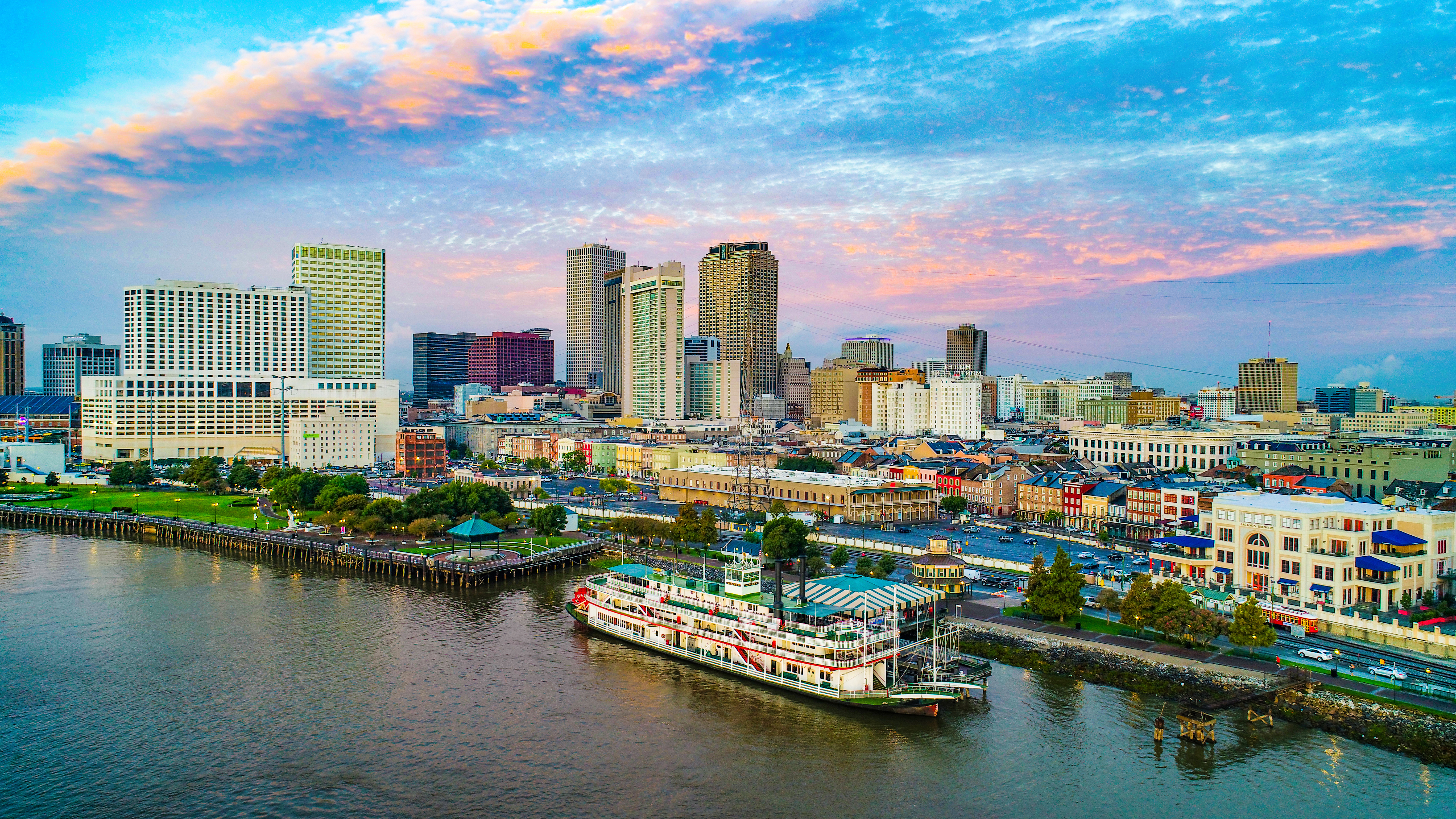 A vibrant cityscape featuring buildings and waterfront, with a steamboat docked in the foreground under a colorful sky.