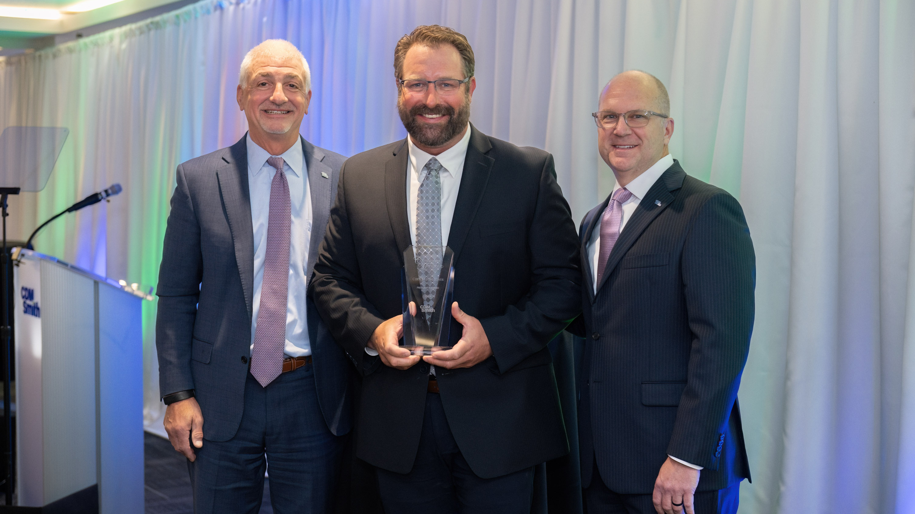 Three men in suits stand on stage, one holding a trophy, with a podium in the background.