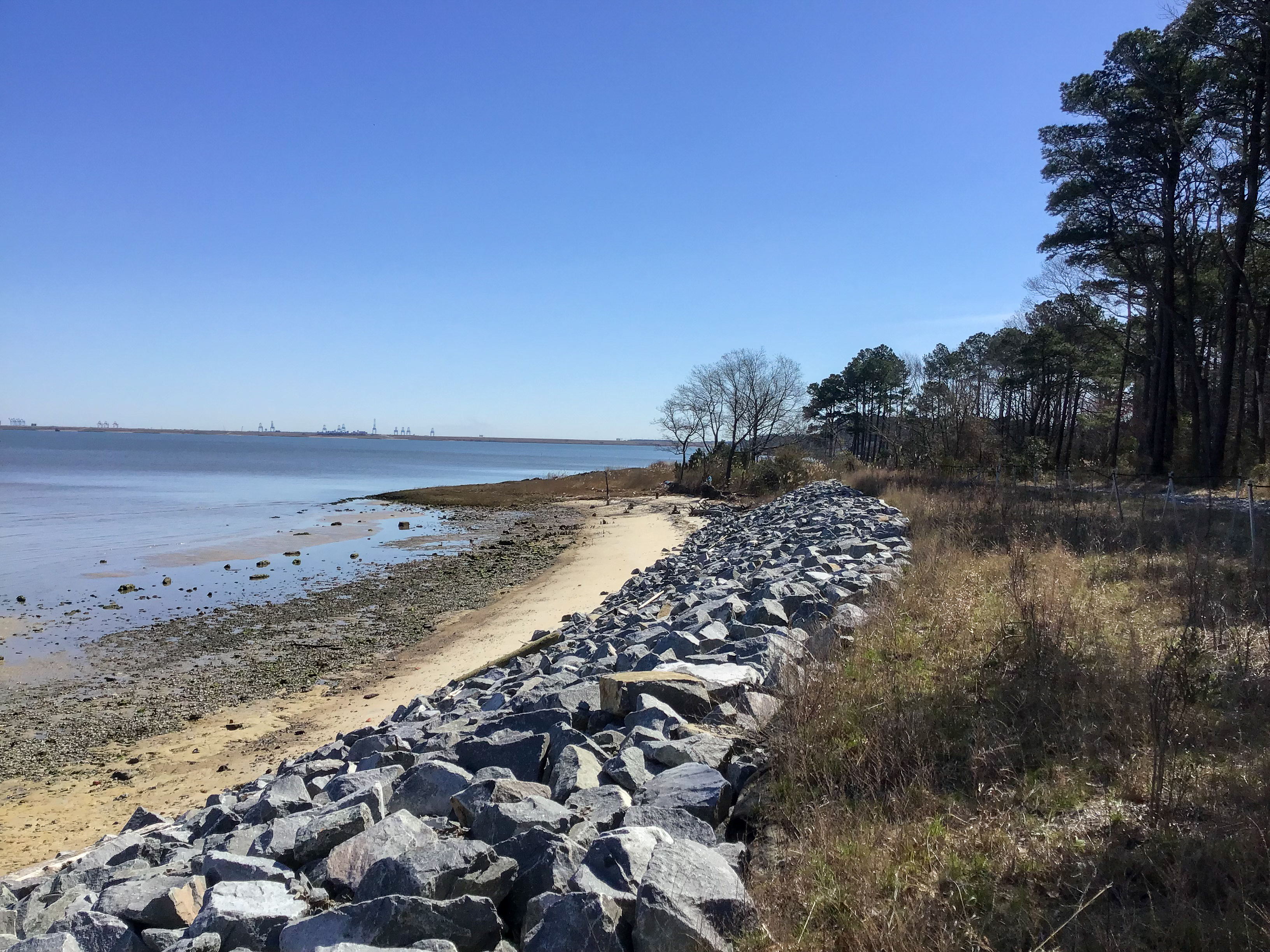 Rocky shoreline with a sandy beach, trees, and distant industrial structures under a blue sky.