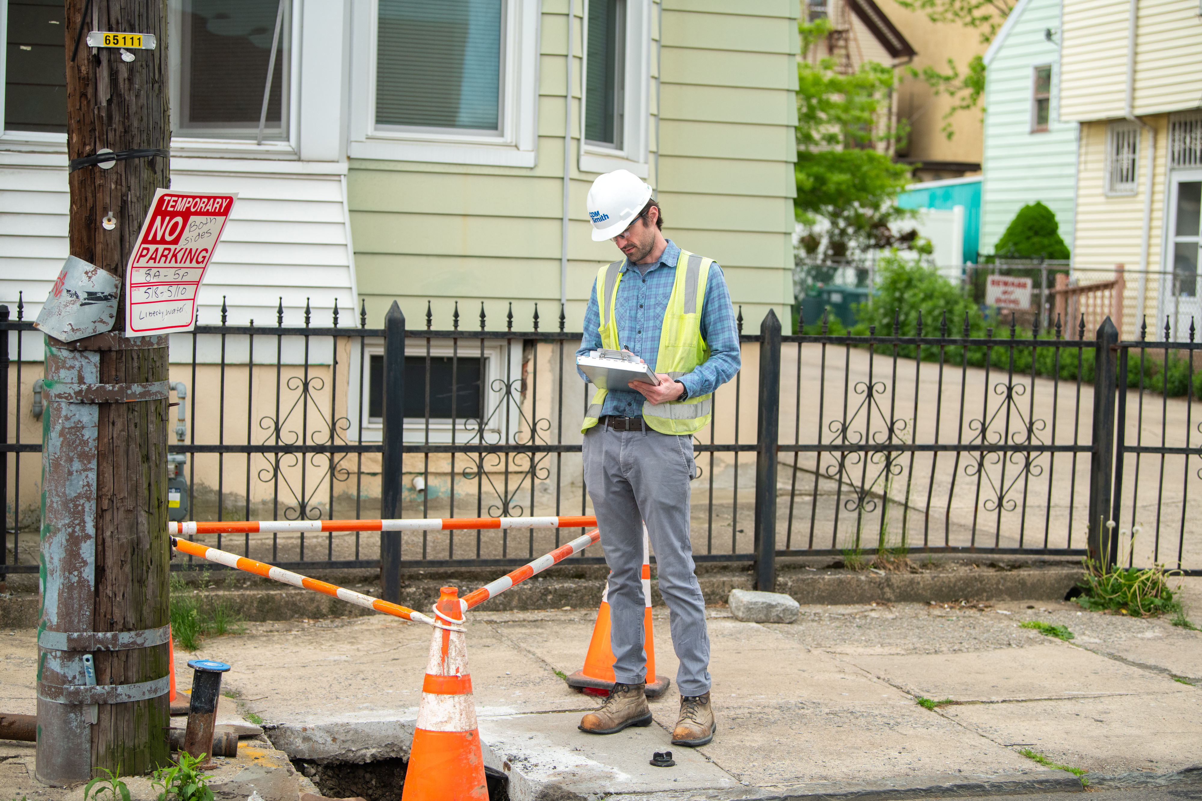 Construction worker in safety gear jotting down notes near a road with traffic cones and no parking sign.