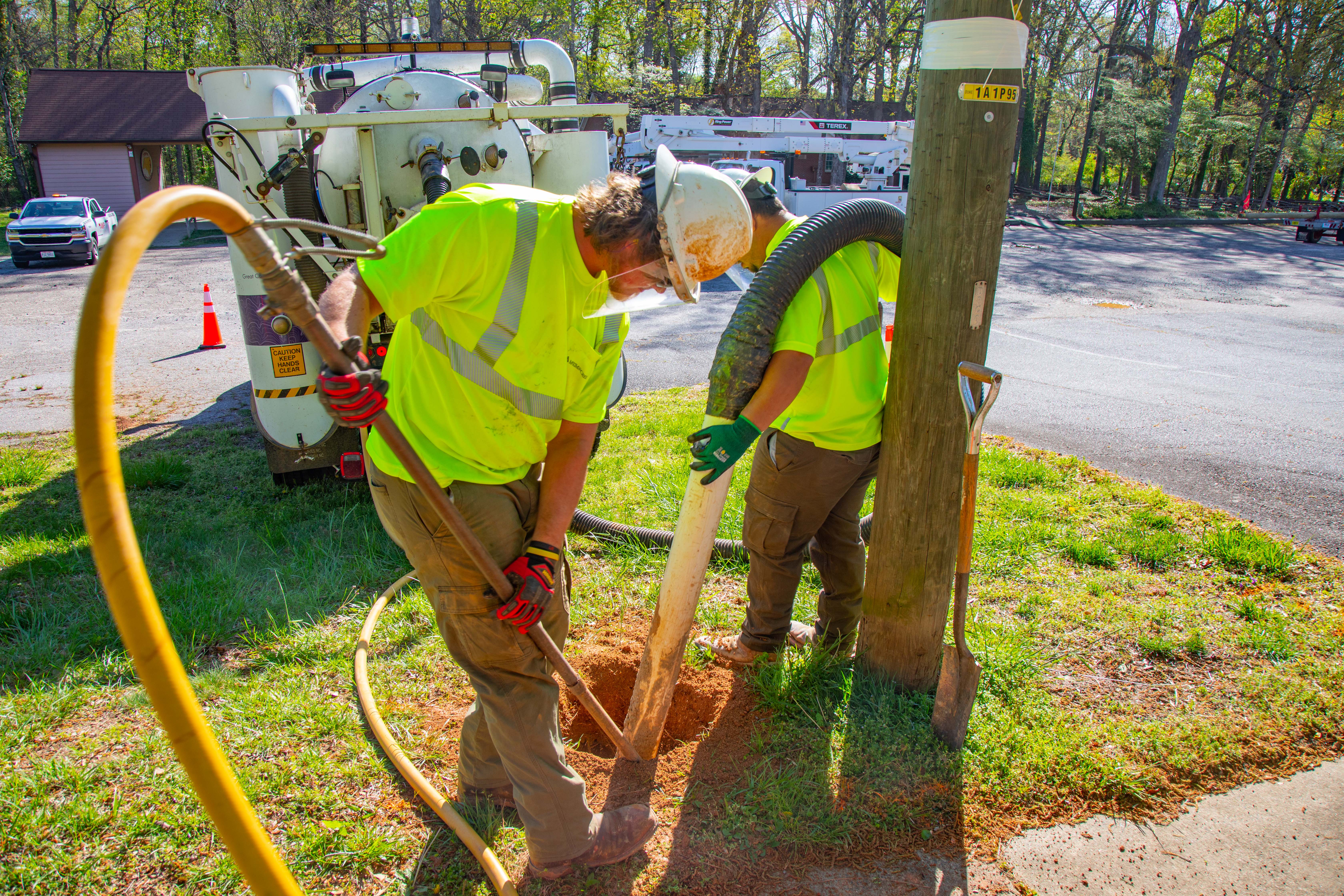 Two workers in high-vis clothing use equipment to inspect a service line near a utility pole.