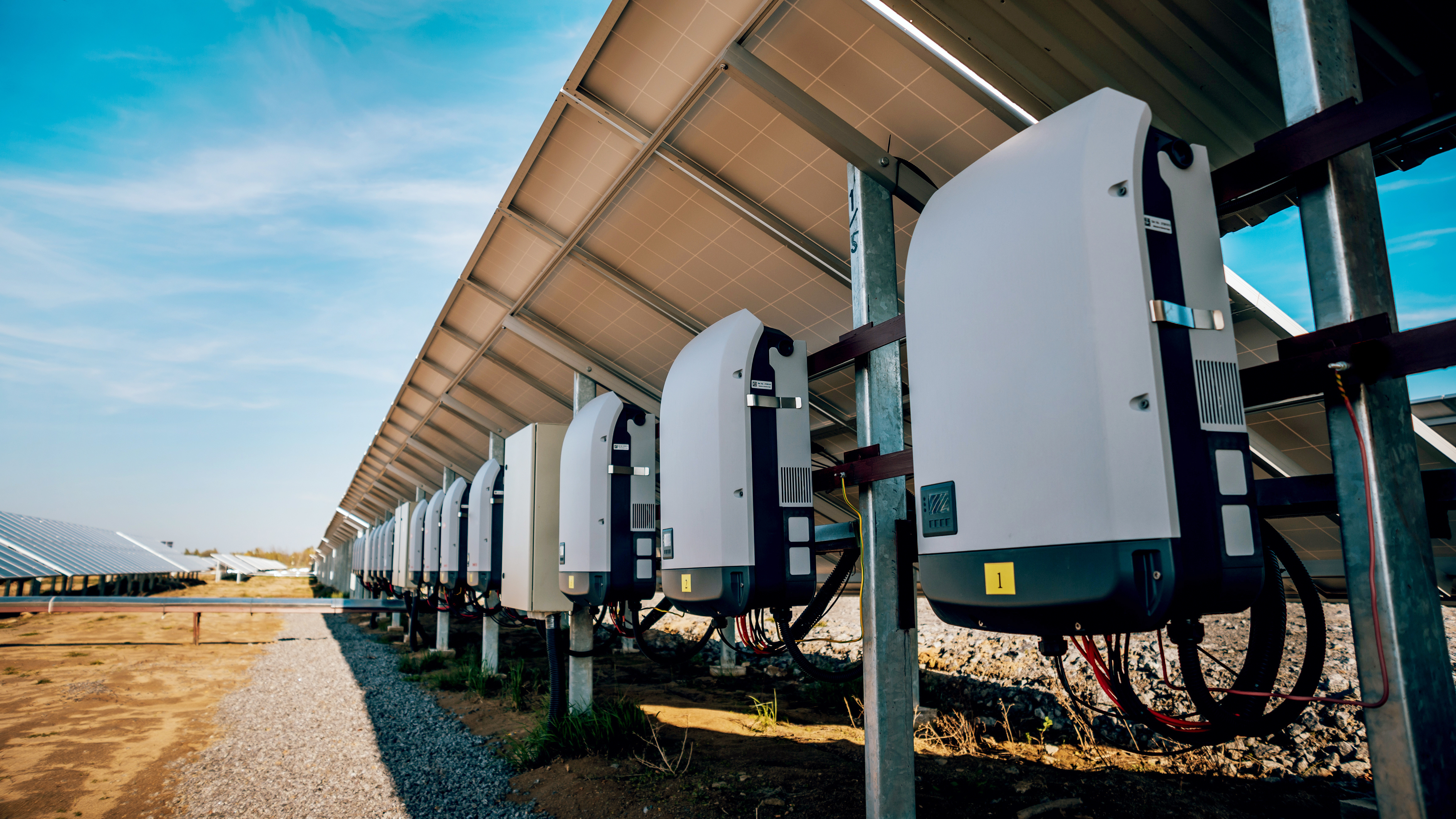 Row of solar panel inverters under solar panels against blue sky.