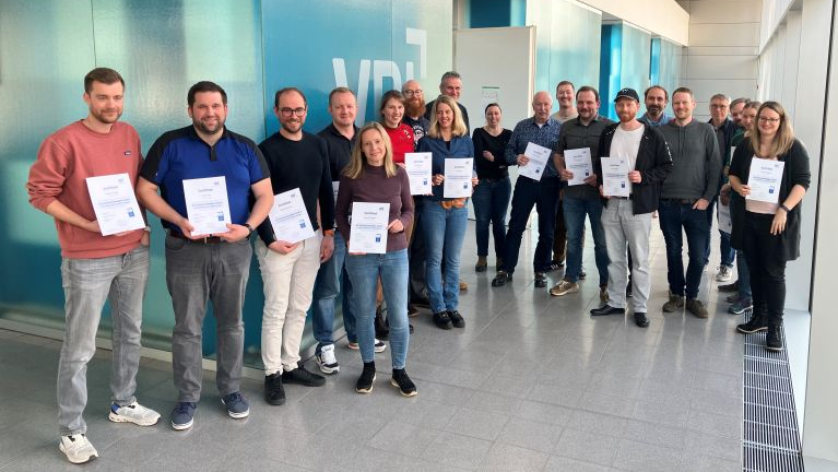Eine Gruppe von Erwachsenen steht in einem Foyer vor einer türkisfarbenen Glaswand und hält jeweils ein Zertifikat in der Hand.