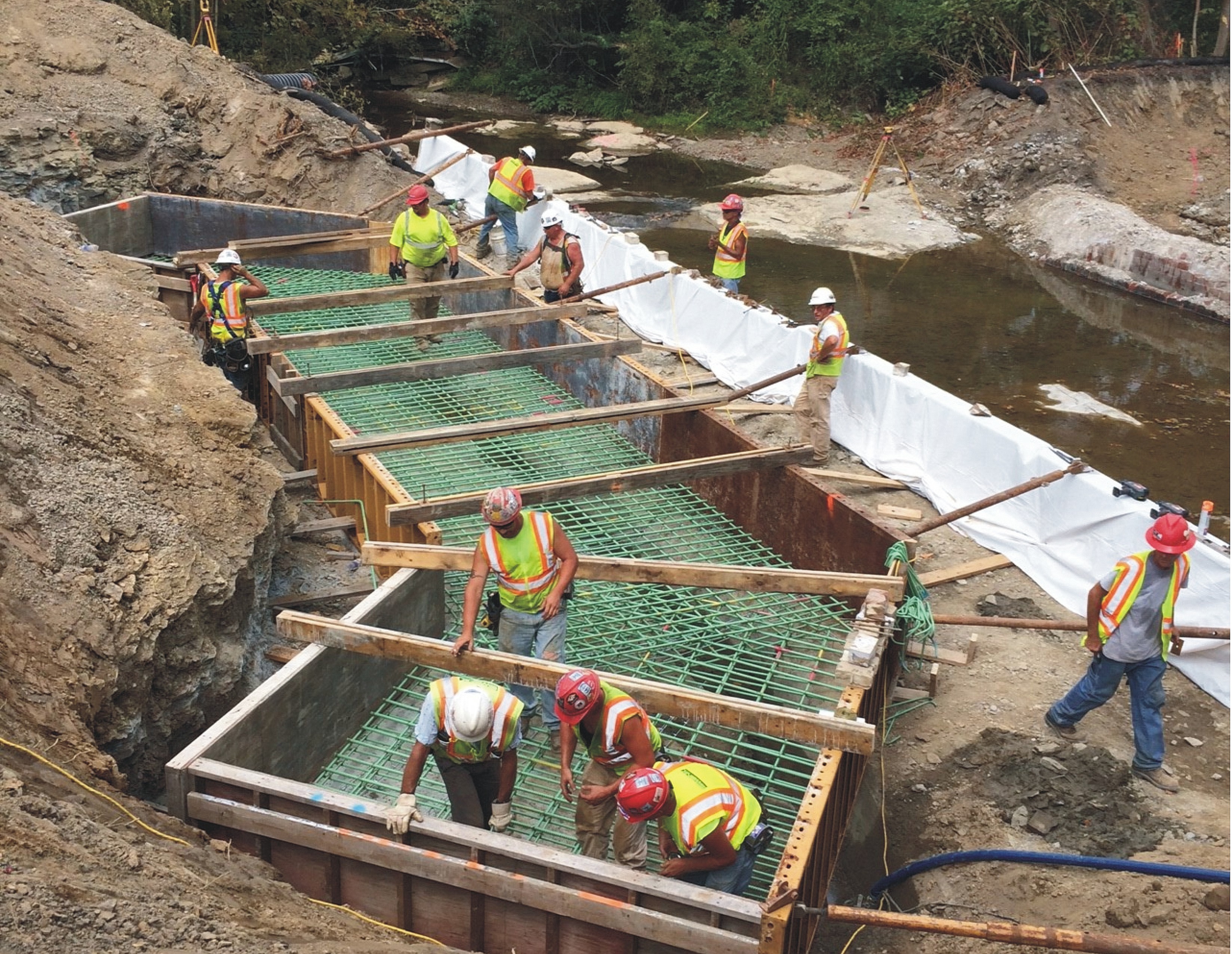 Construction workers building bridge foundations by a river, using wooden frames and rebar, surrounded by earth and rock.