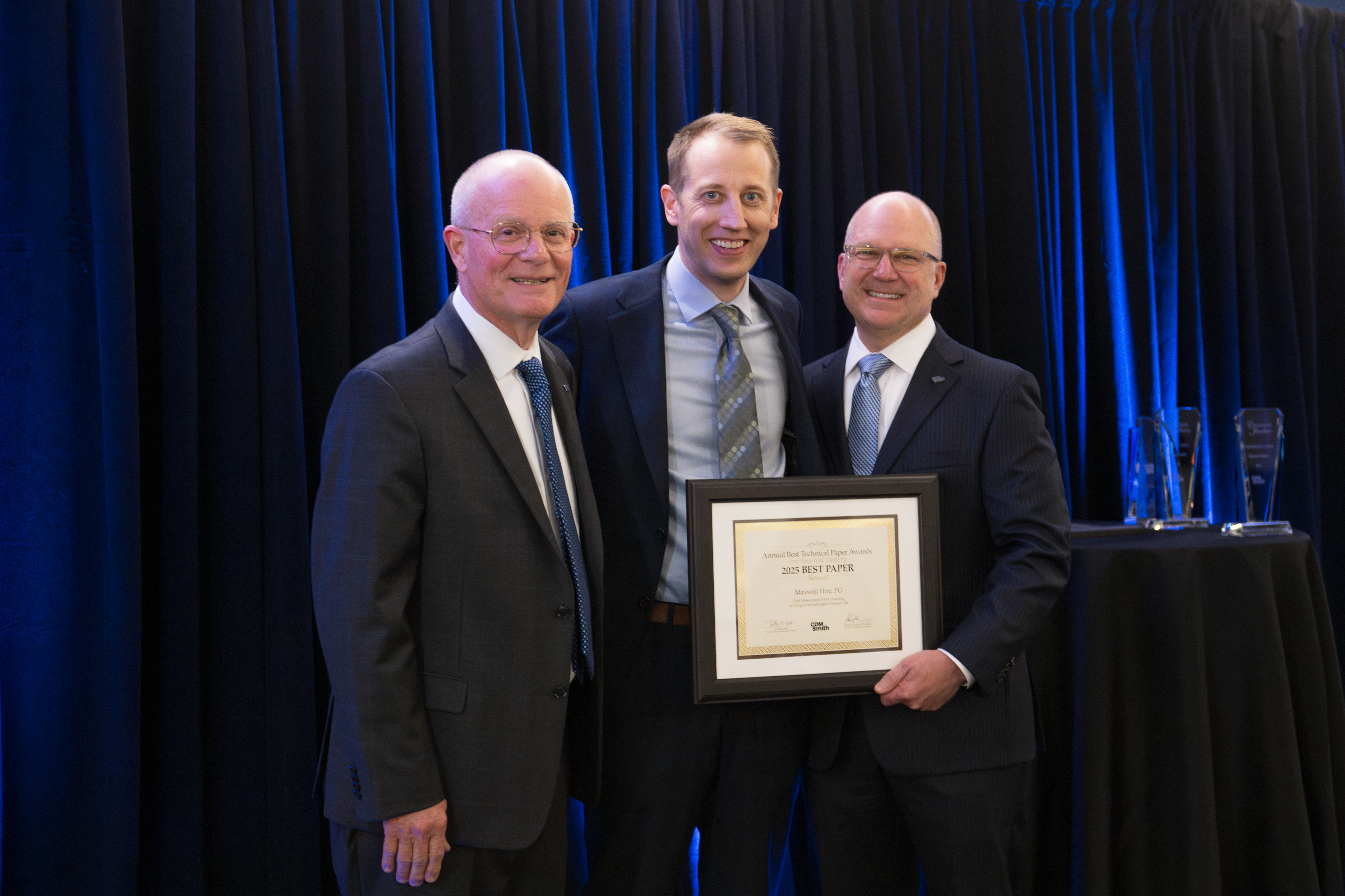 Three individuals in business attire posing for a photo, one holding an award certificate, in front of a blue curtain backdrop.