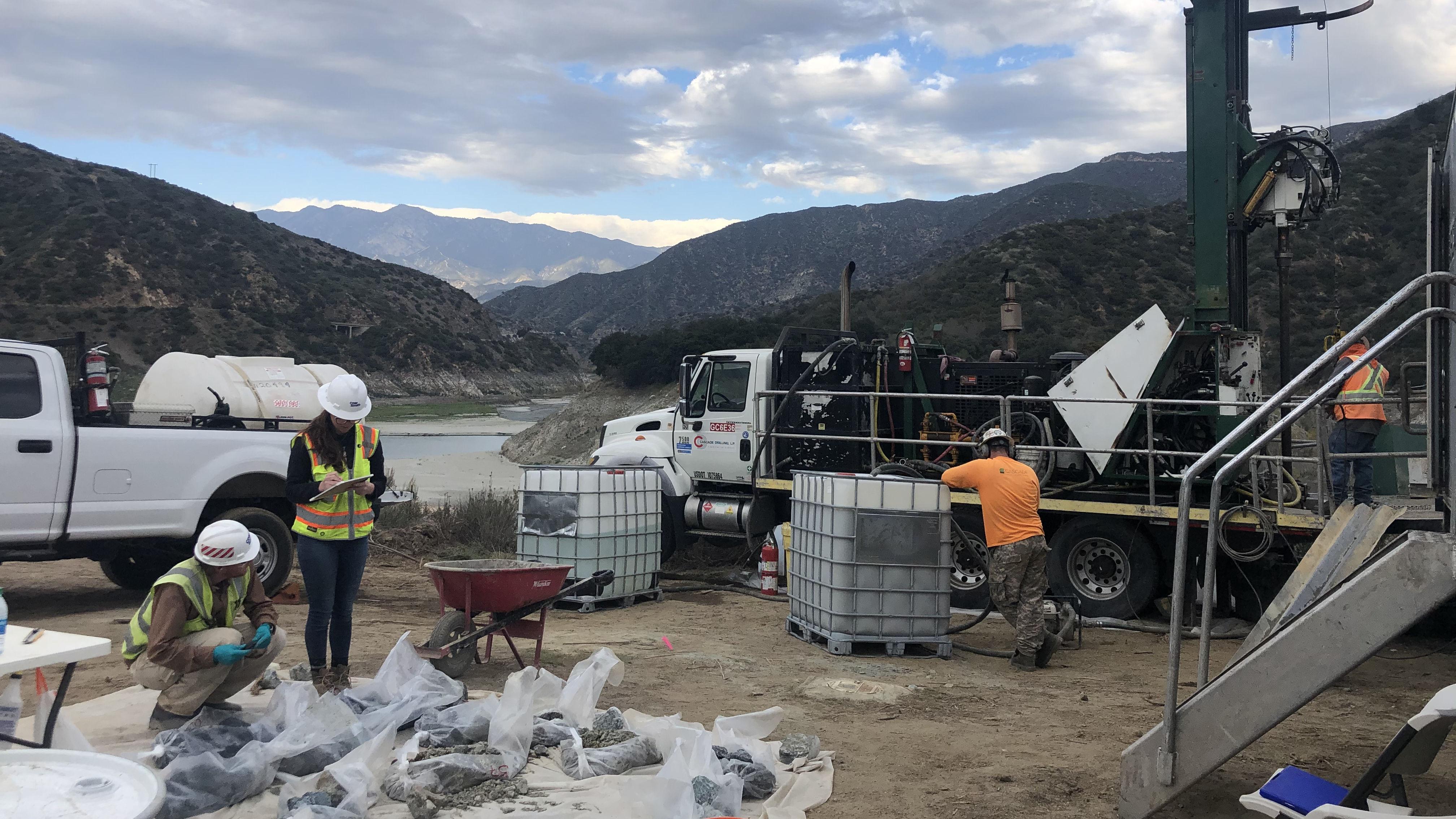 Workers in safety gear at a drilling site near mountains, checking equipment and samples.