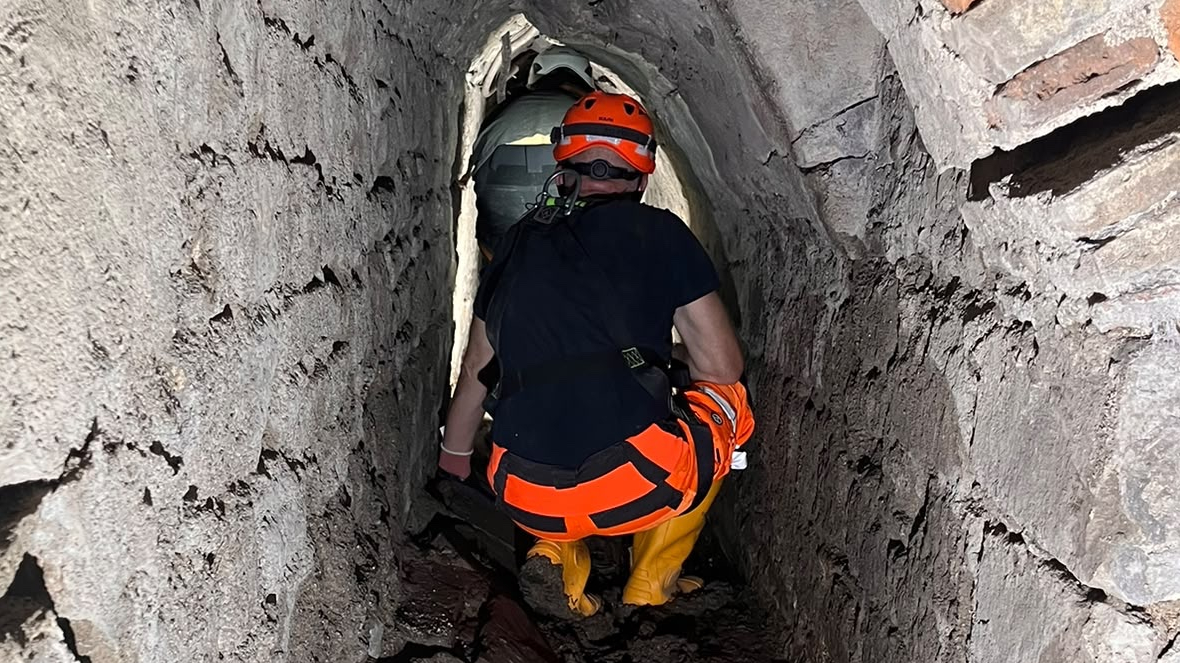 Worker in an orange helmet crouches in a narrow, damaged brick tunnel with a muddy floor.