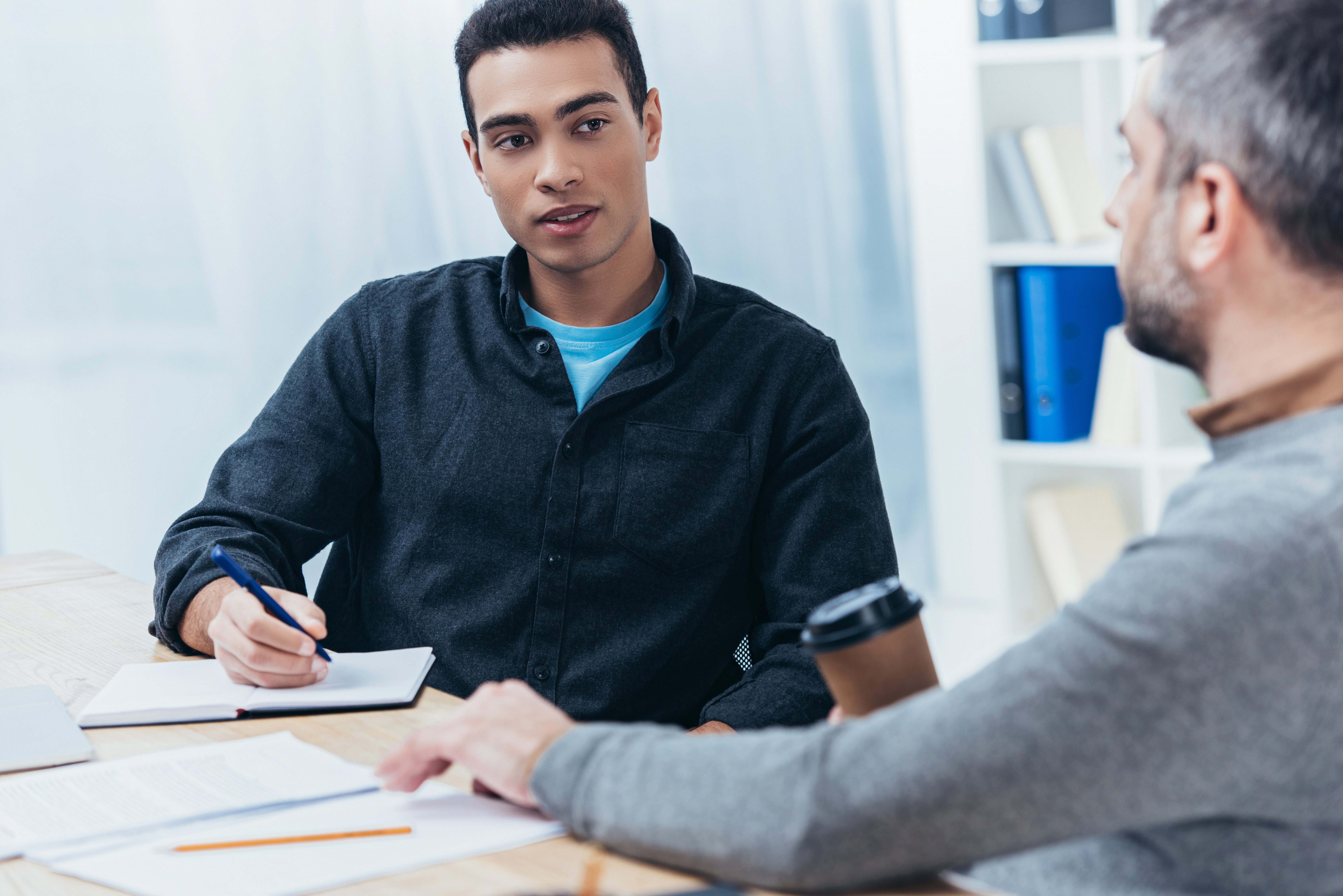 Two individuals seated at a table engaged in a discussion, with notebooks and papers on the surface.