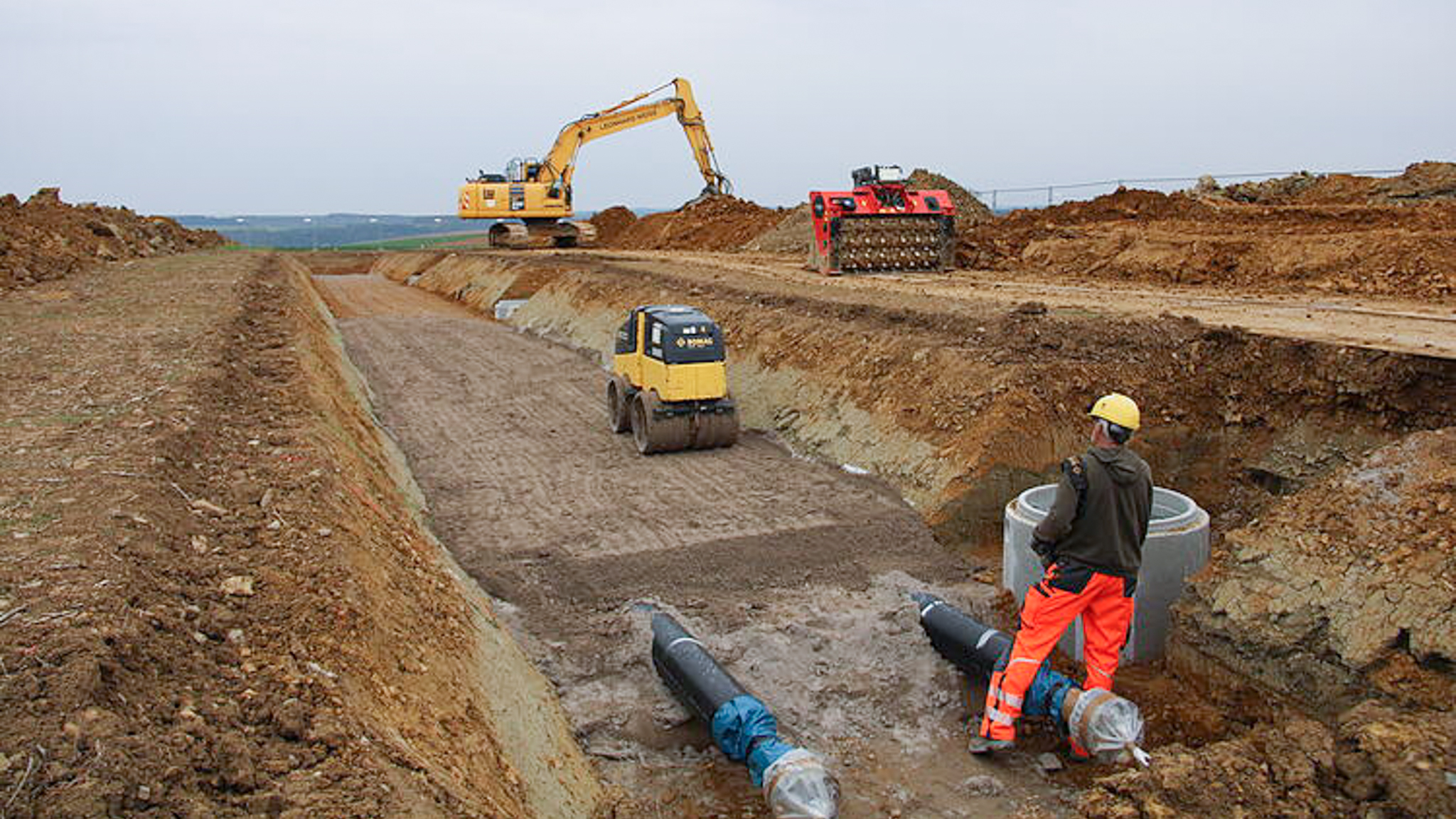 Construction site with an excavator in a trench, worker in safety gear, and large pipes visible.