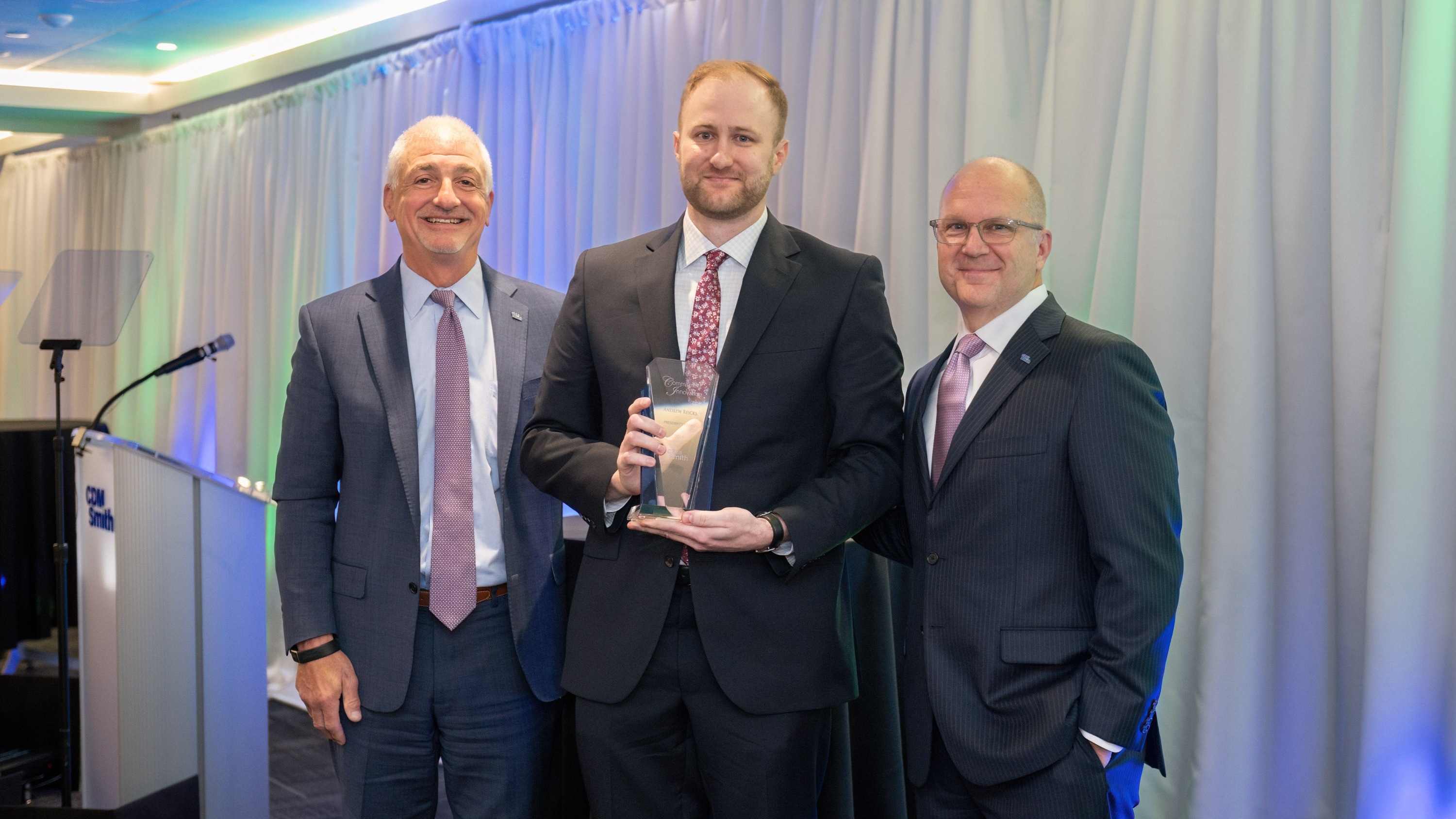 Three men in suits, one holding an award, standing in front of a curtain at an event.