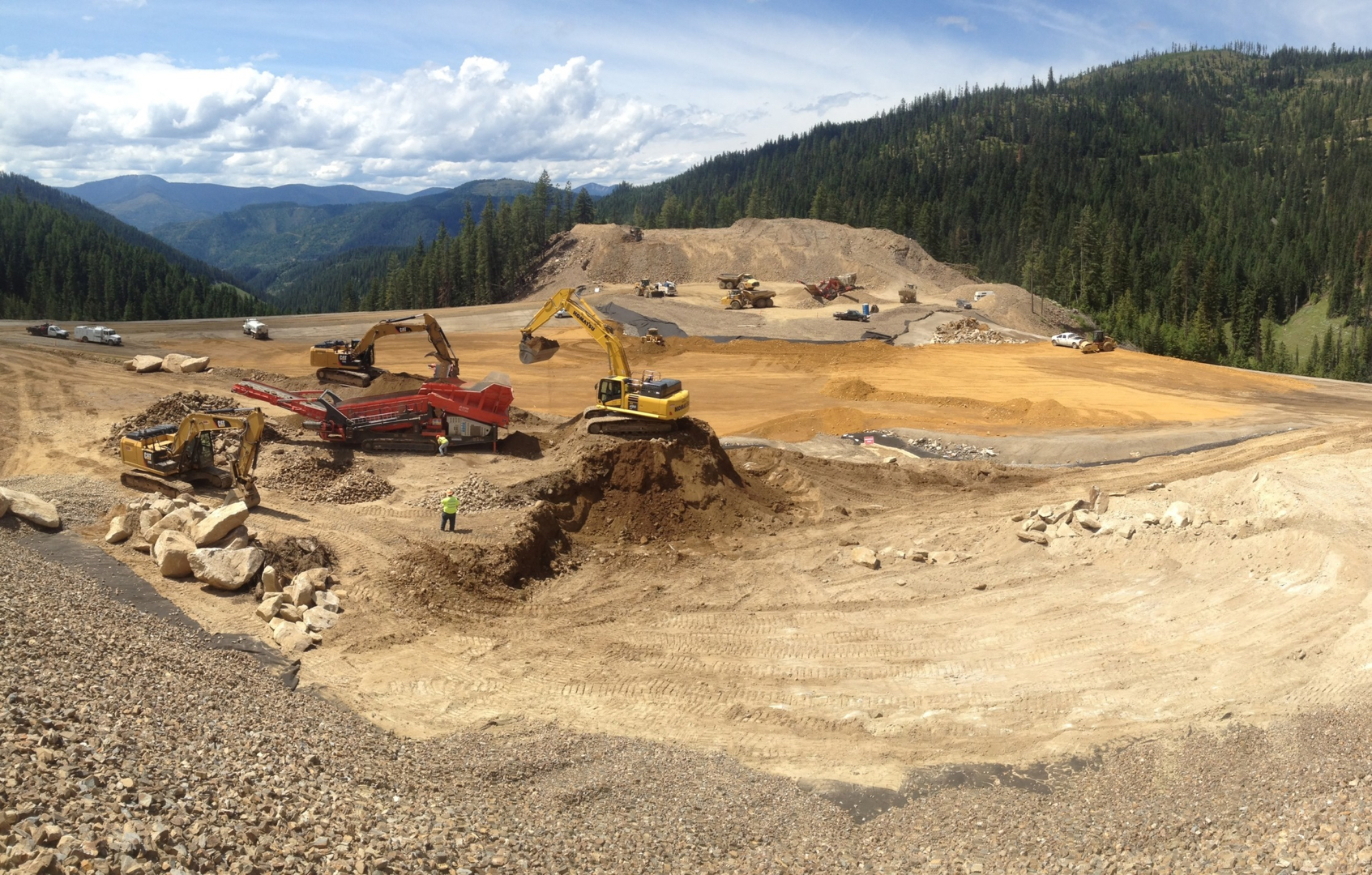 Construction machinery working on a large dirt site surrounded by forested hills and mountains.