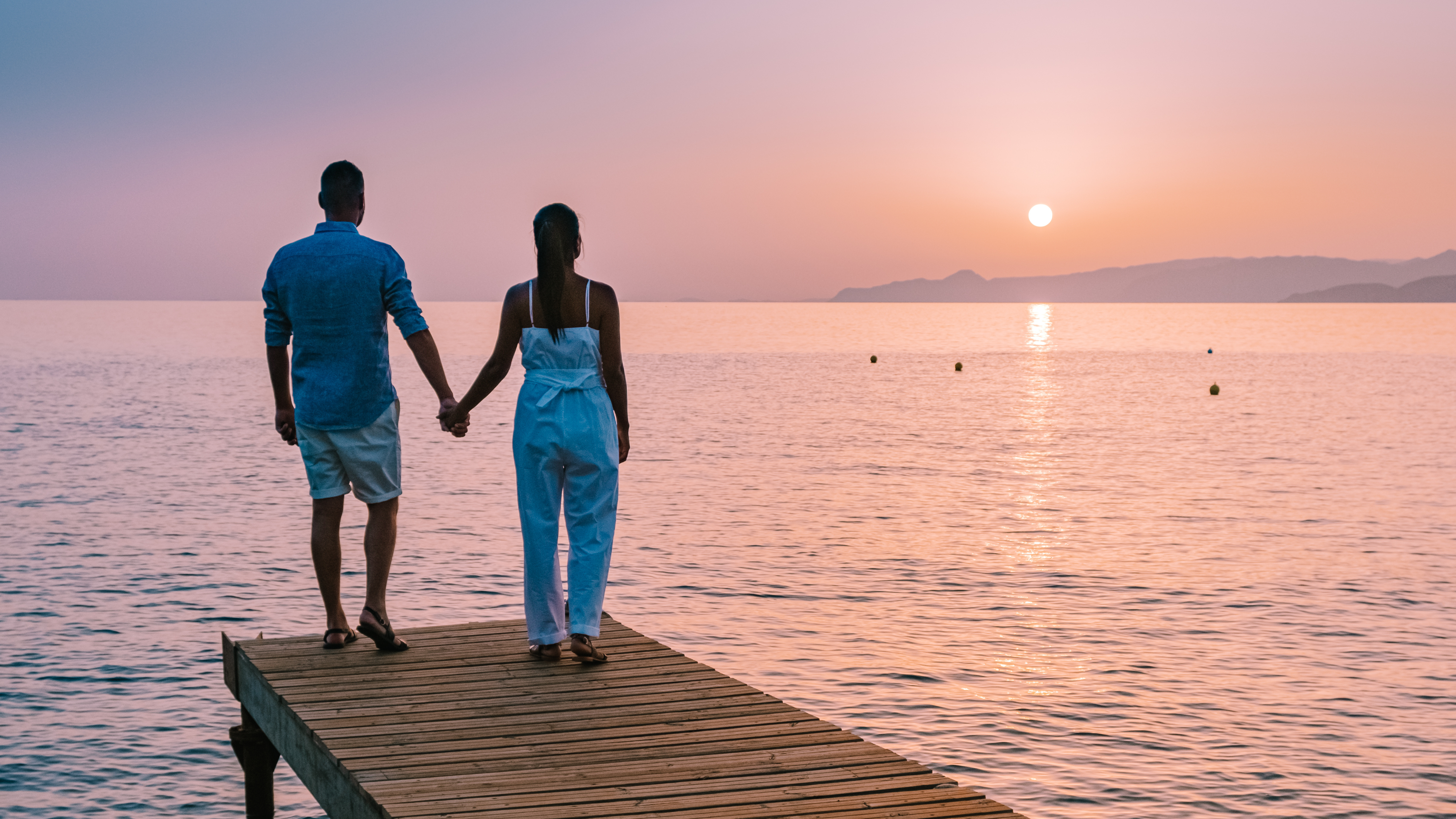 A couple holding hands on a dock at sunset, overlooking the ocean.