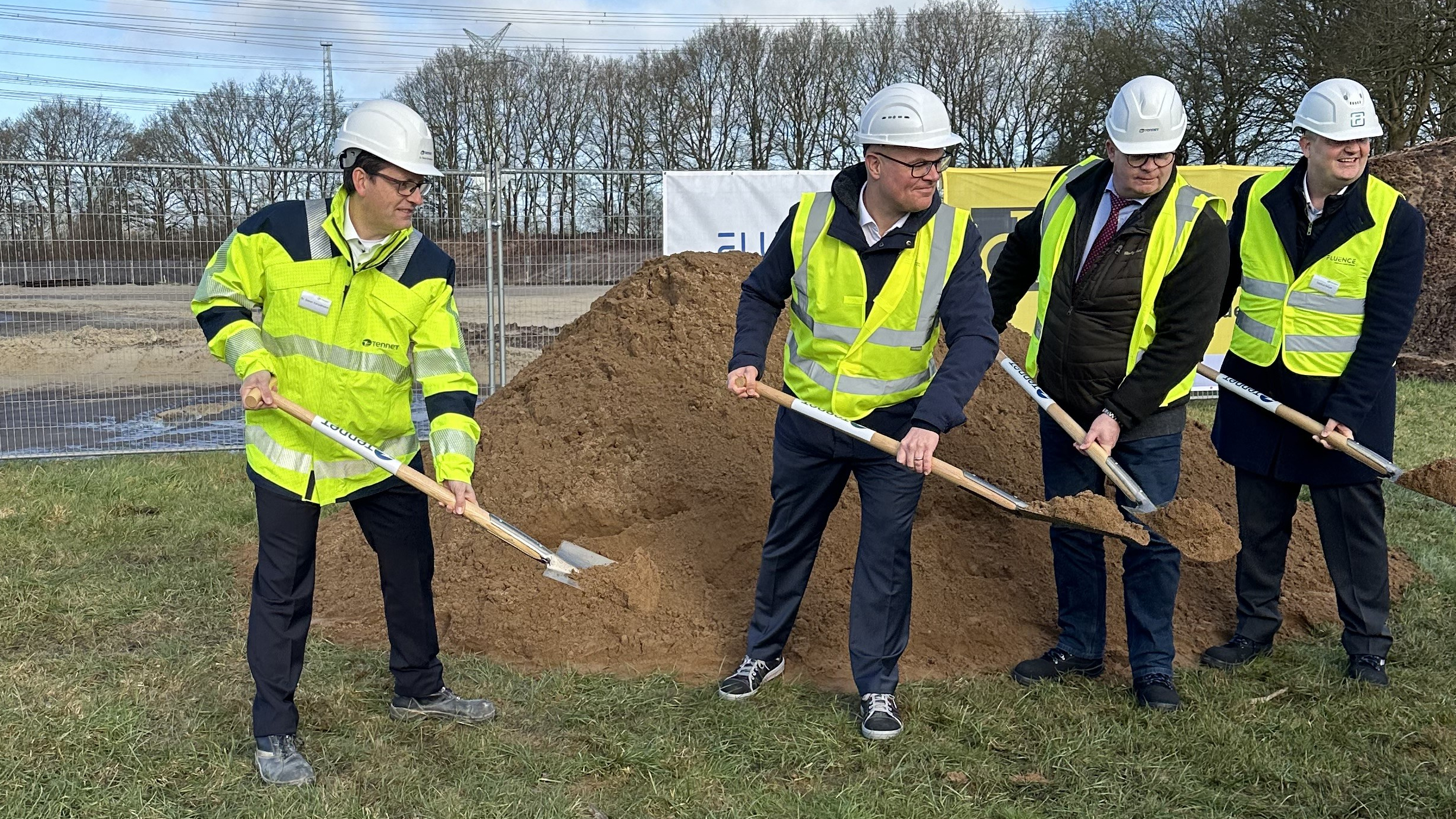 Four people in safety gear perform a ceremonial groundbreaking with shovels in front of a dirt mound near a TenneT banner.