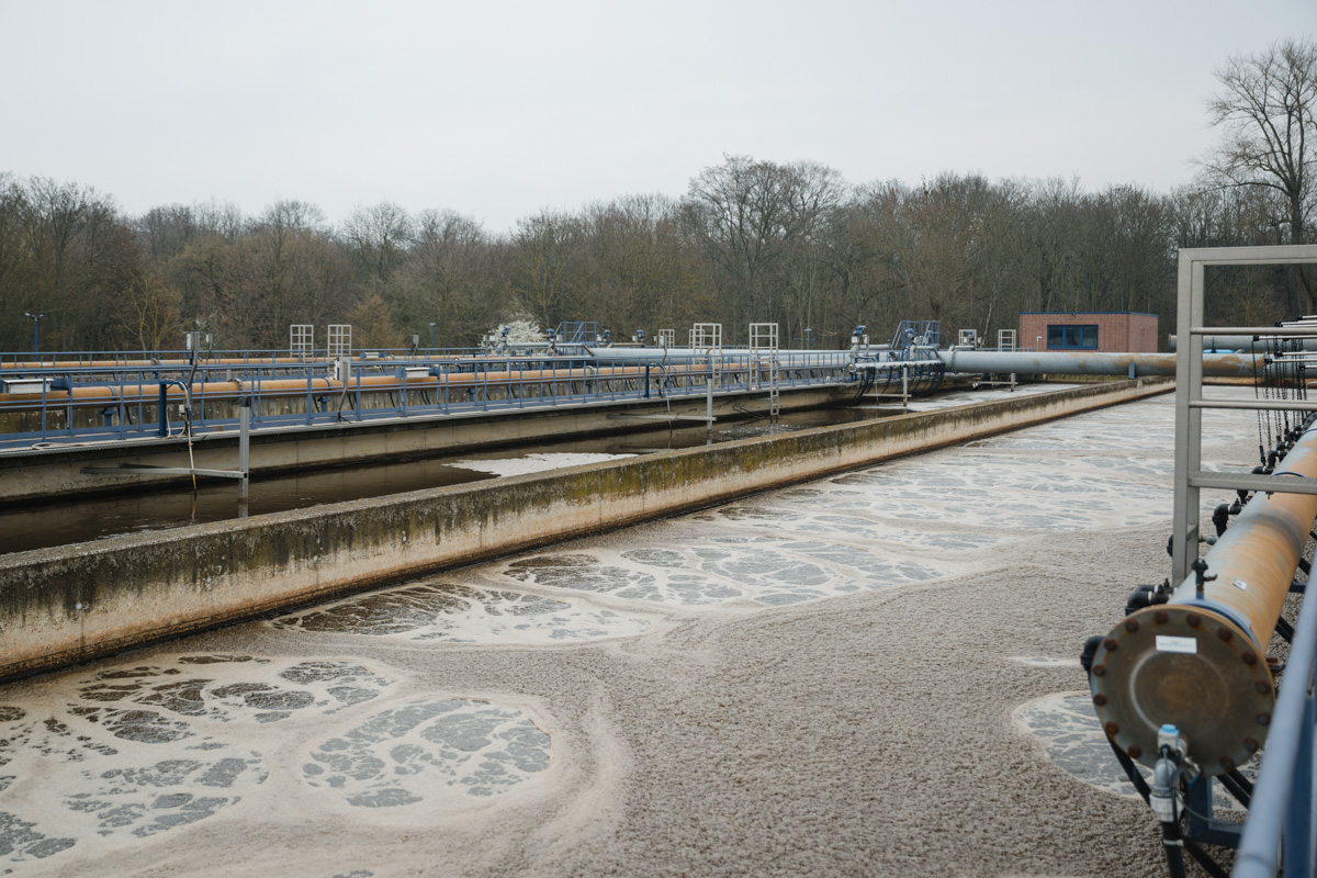 A wastewater treatment facility with large tanks processing water. Pipes and equipment are visible, surrounded by trees.