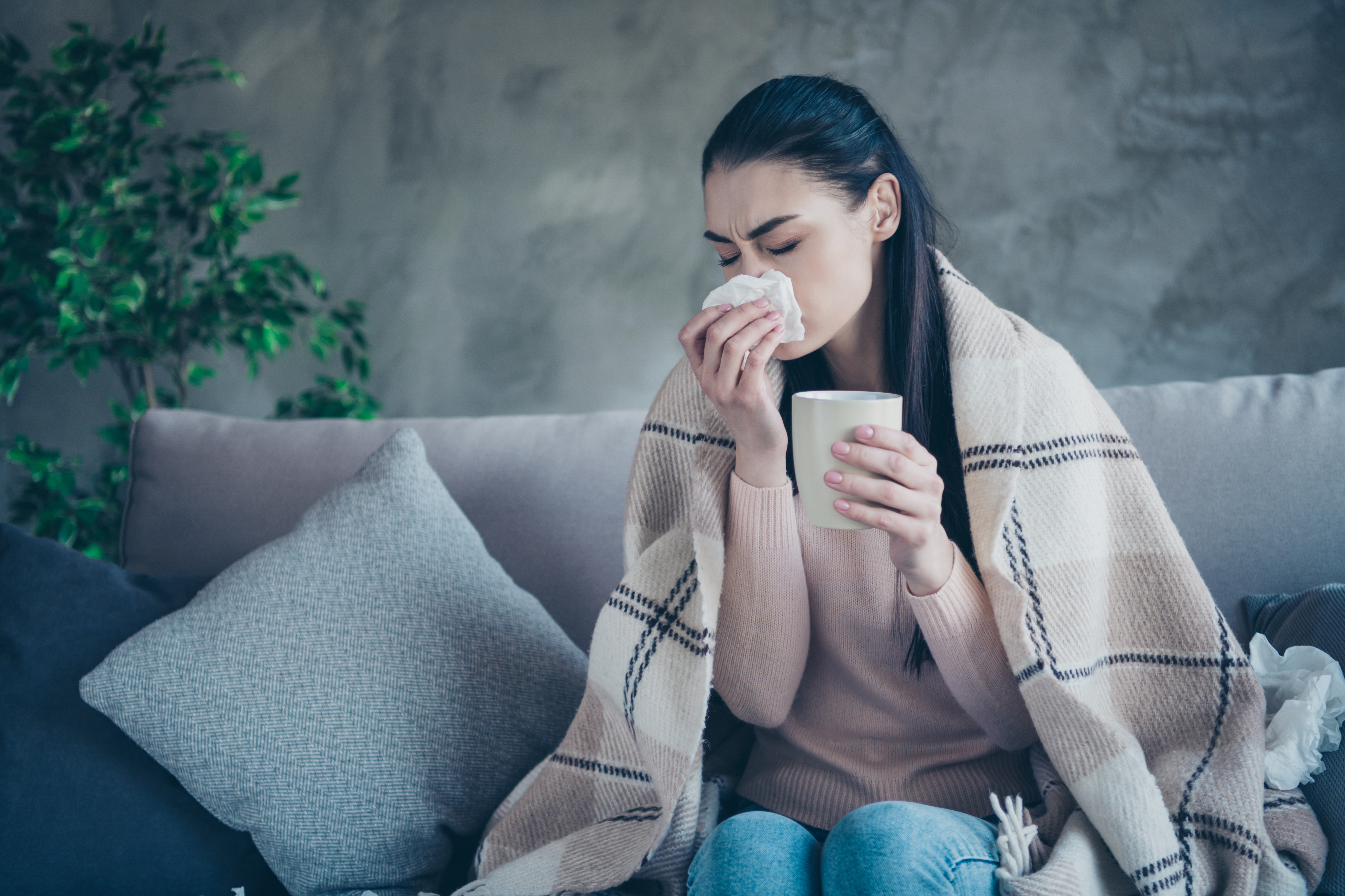 A person wrapped in a blanket sitting on a couch, holding a cup, with a tissue box nearby.