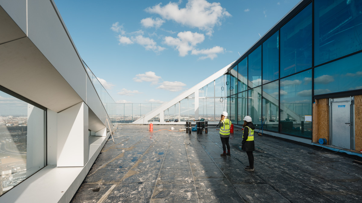 A rooftop view featuring modern architecture, with two construction workers wearing safety gear standing near a glass facade.