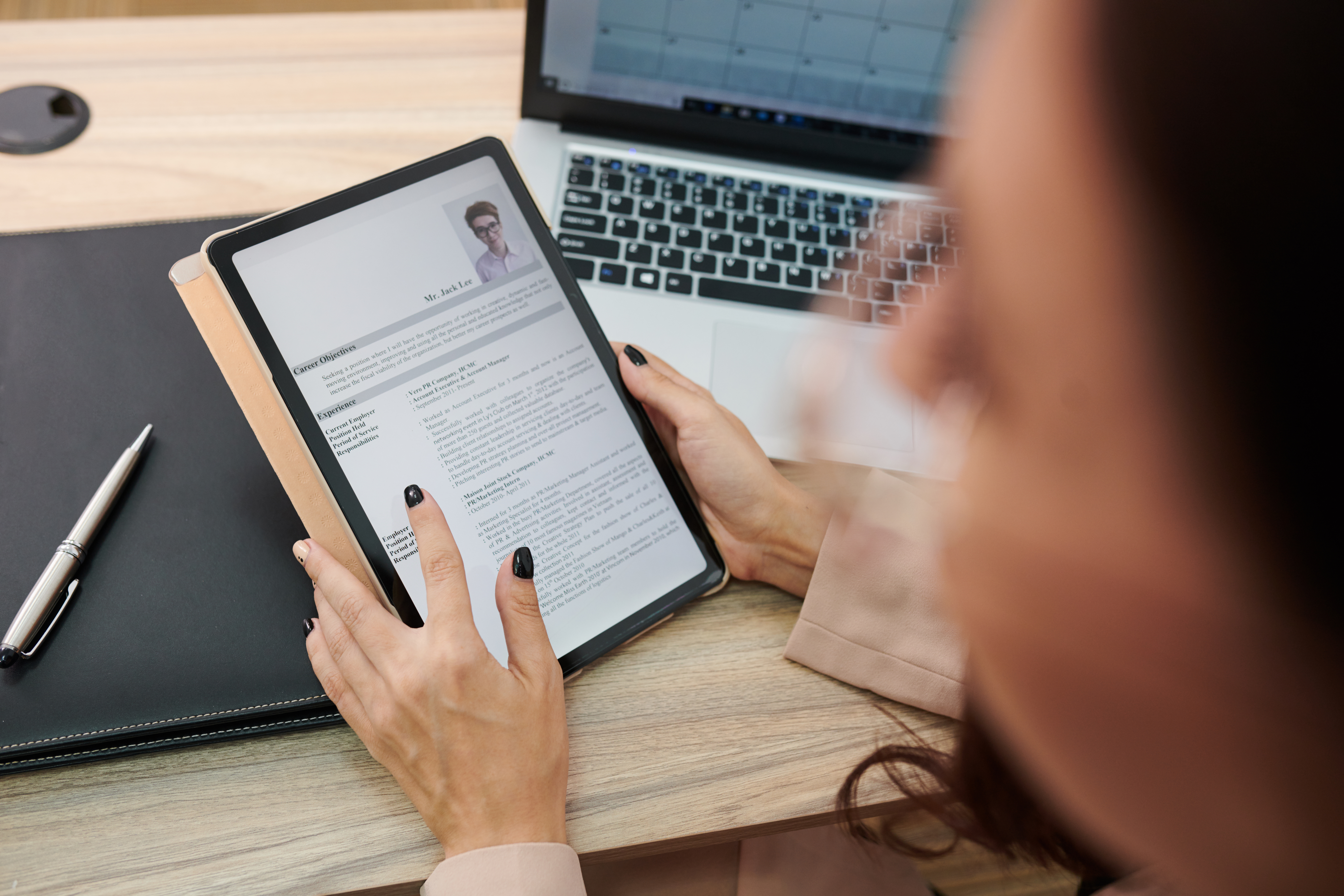 Person holding a tablet displaying a professional resume at a desk with a laptop and pen nearby.