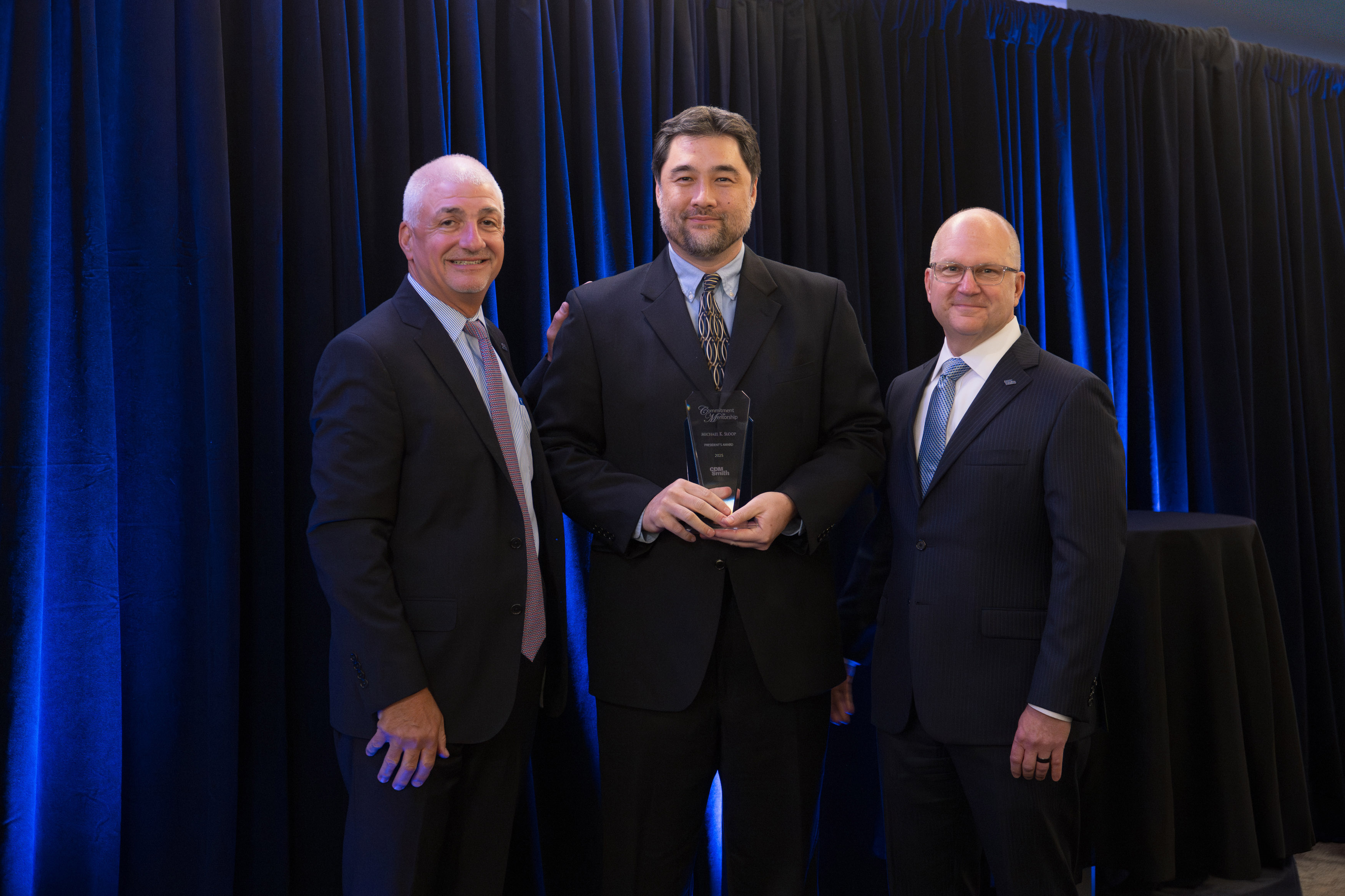 Three individuals in formal attire standing in front of a blue curtain, one holding a glass award.