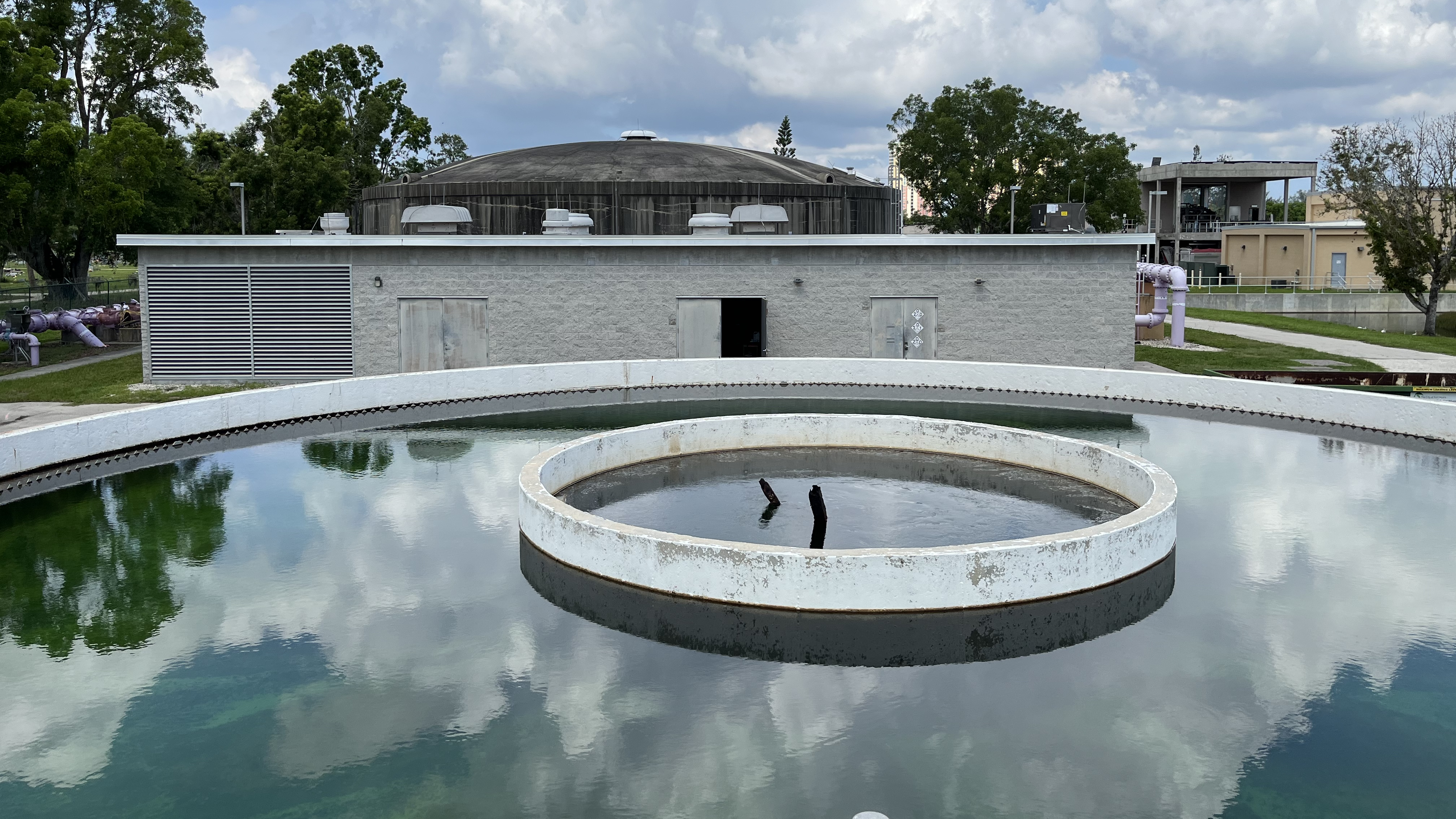 Water treatment facility with circular tanks and a domed building, surrounded by trees under a cloudy sky.