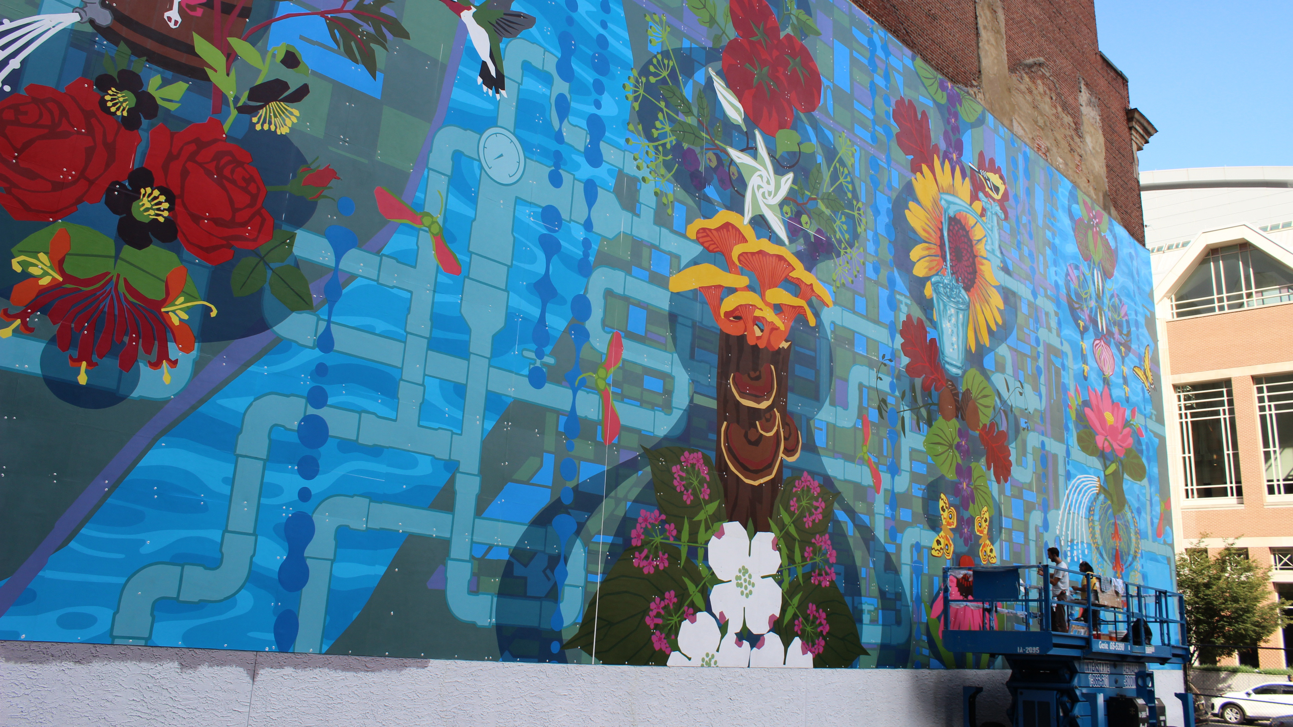 Colorful mural of nature with flowers, birds, and pipes on a brick wall; people working from a lift below.