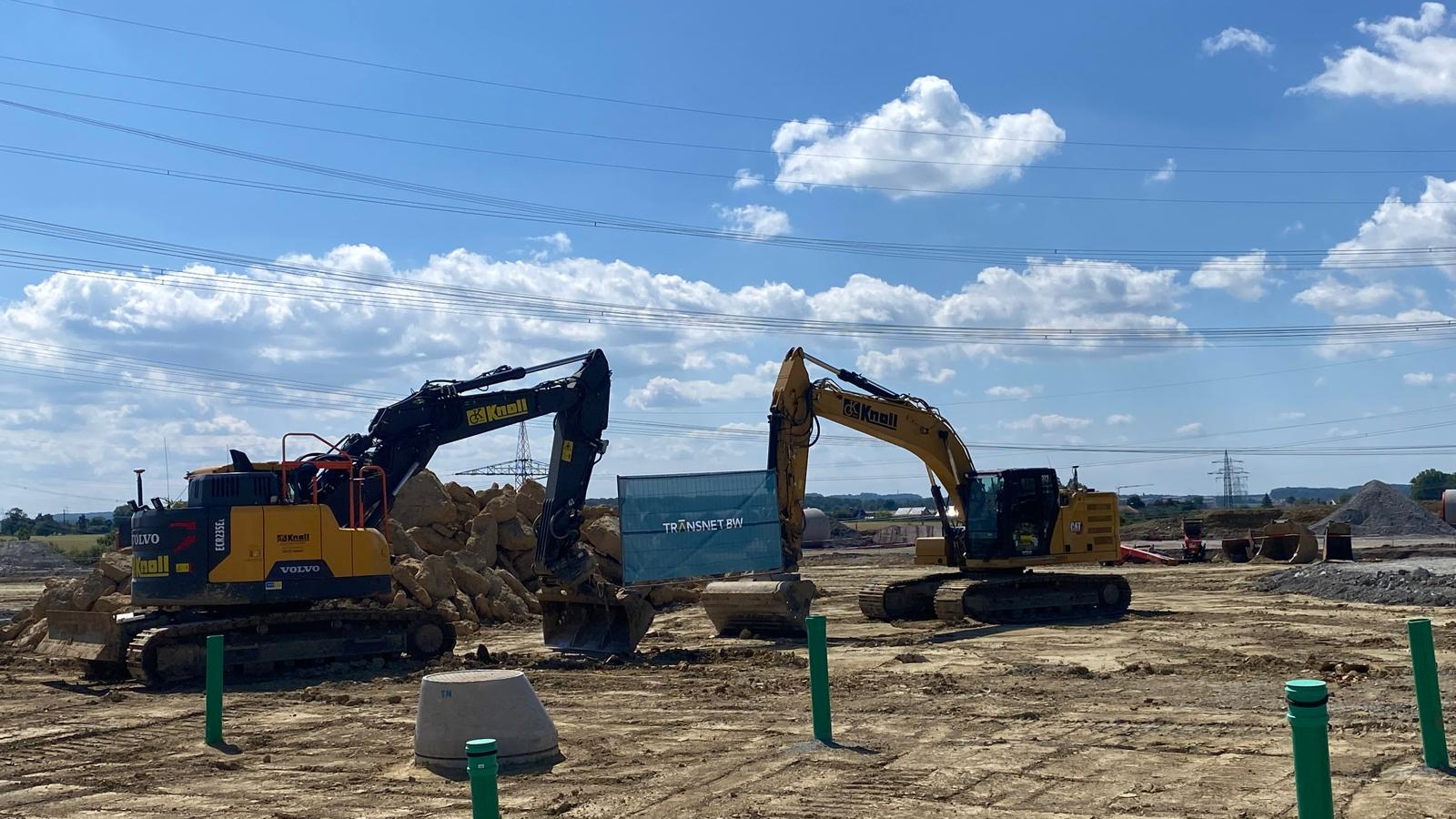 Two excavators digging at a construction site under a blue sky with clouds.