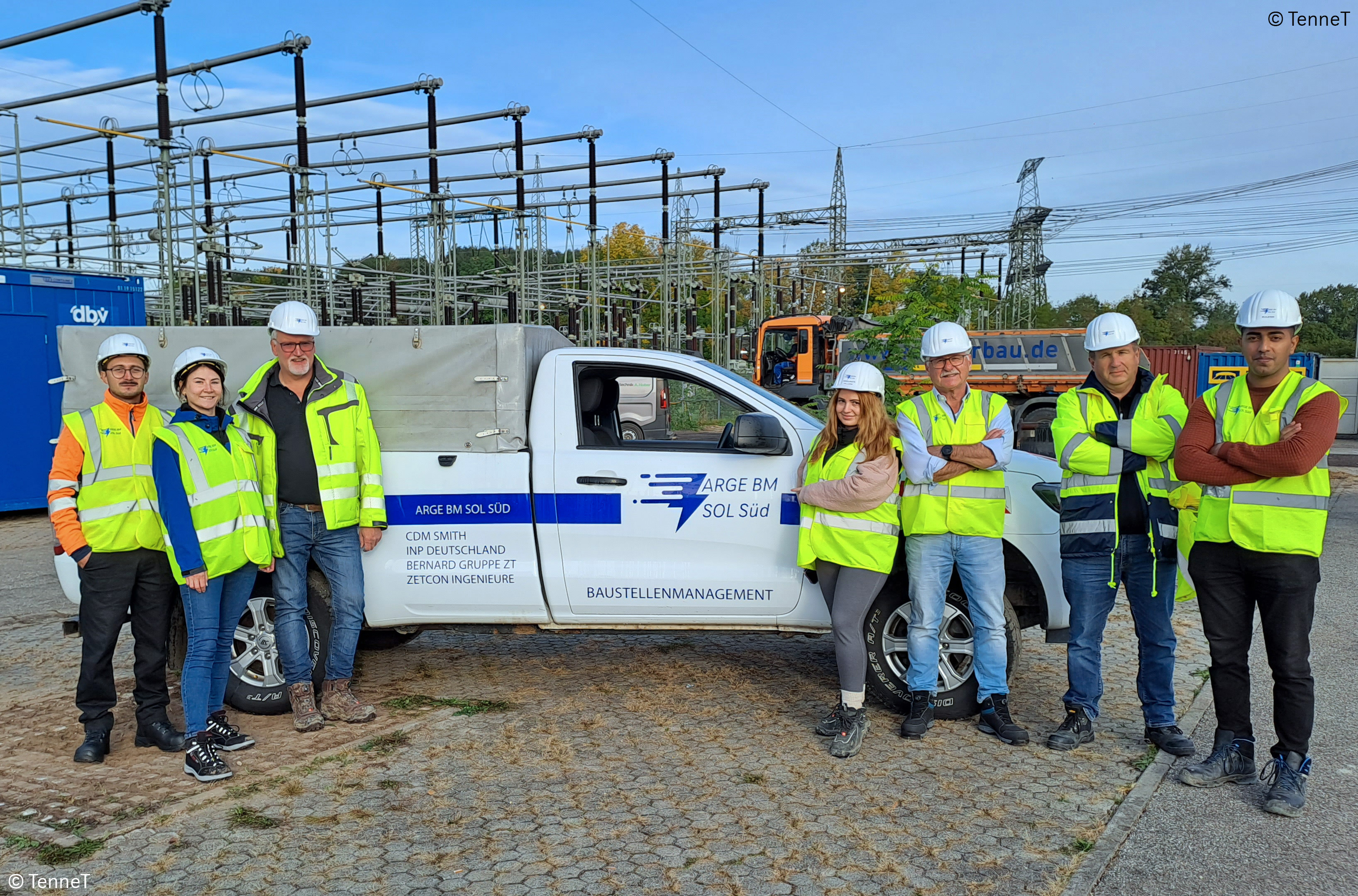 Workers in safety vests and helmets stand by a white utility truck at a construction site.