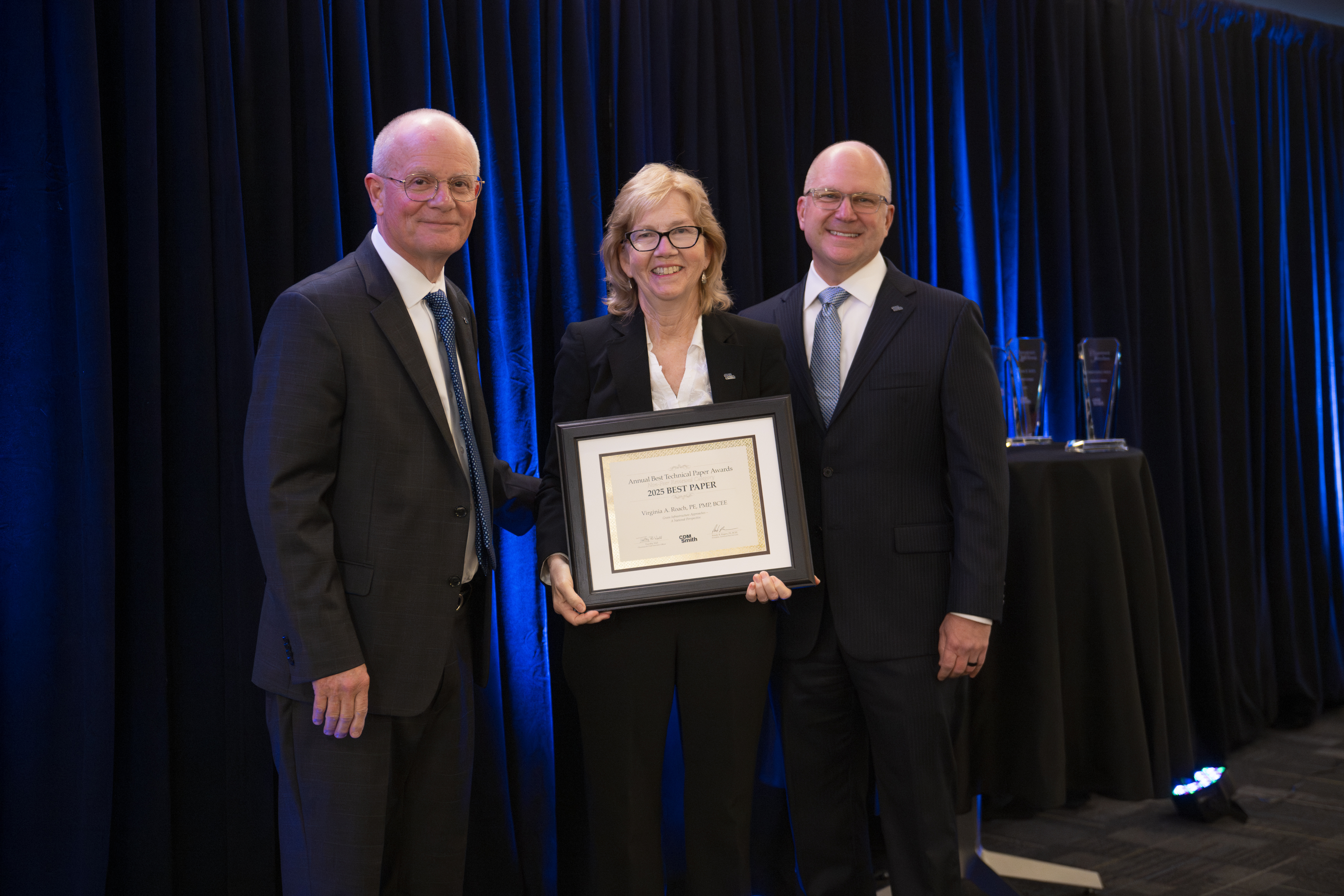 Three individuals standing together on a stage, one holding a framed certificate, with dark curtains and a table in the background.