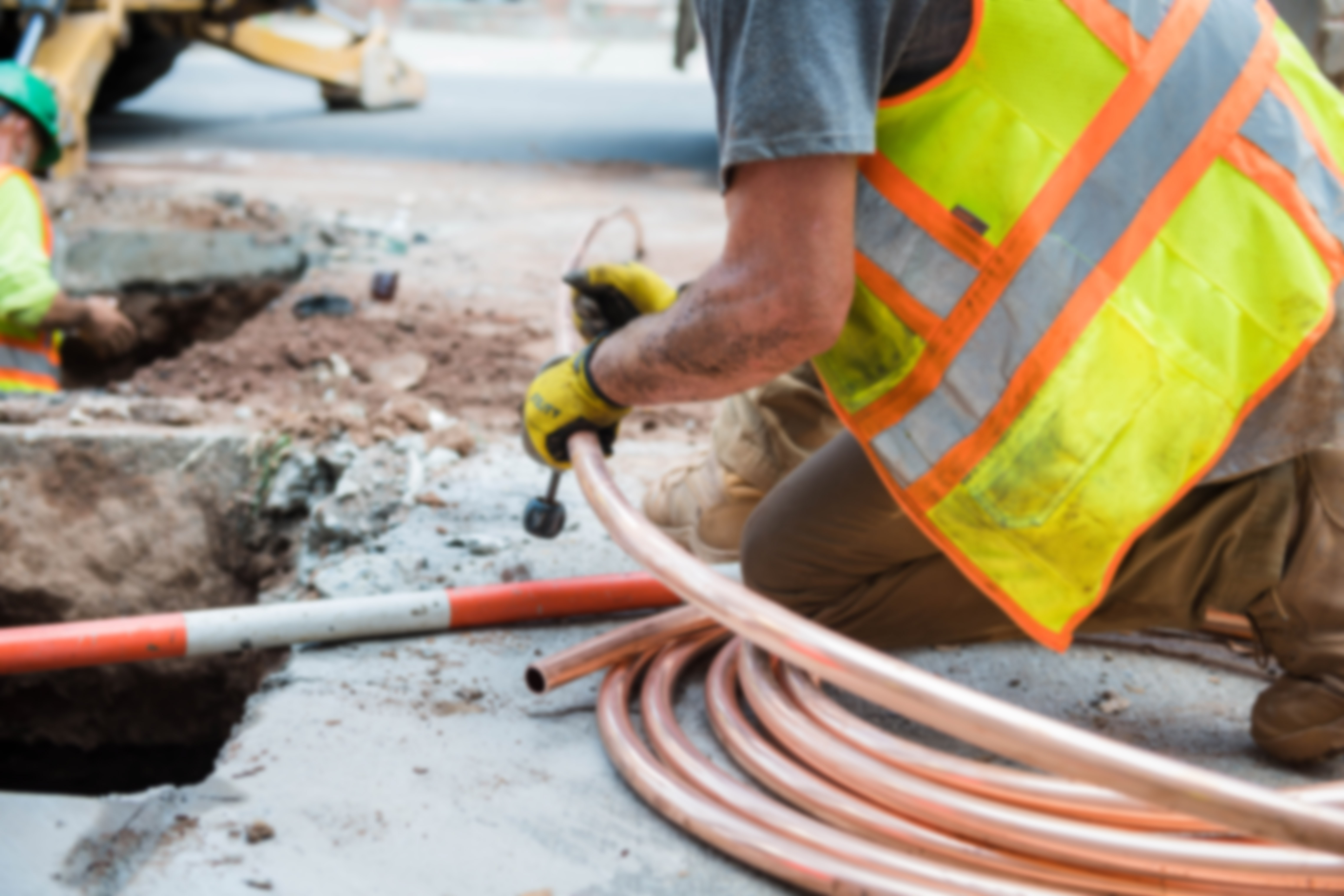 Construction worker kneeling, handling copper tubing next to a trench, wearing a neon safety vest and gloves.