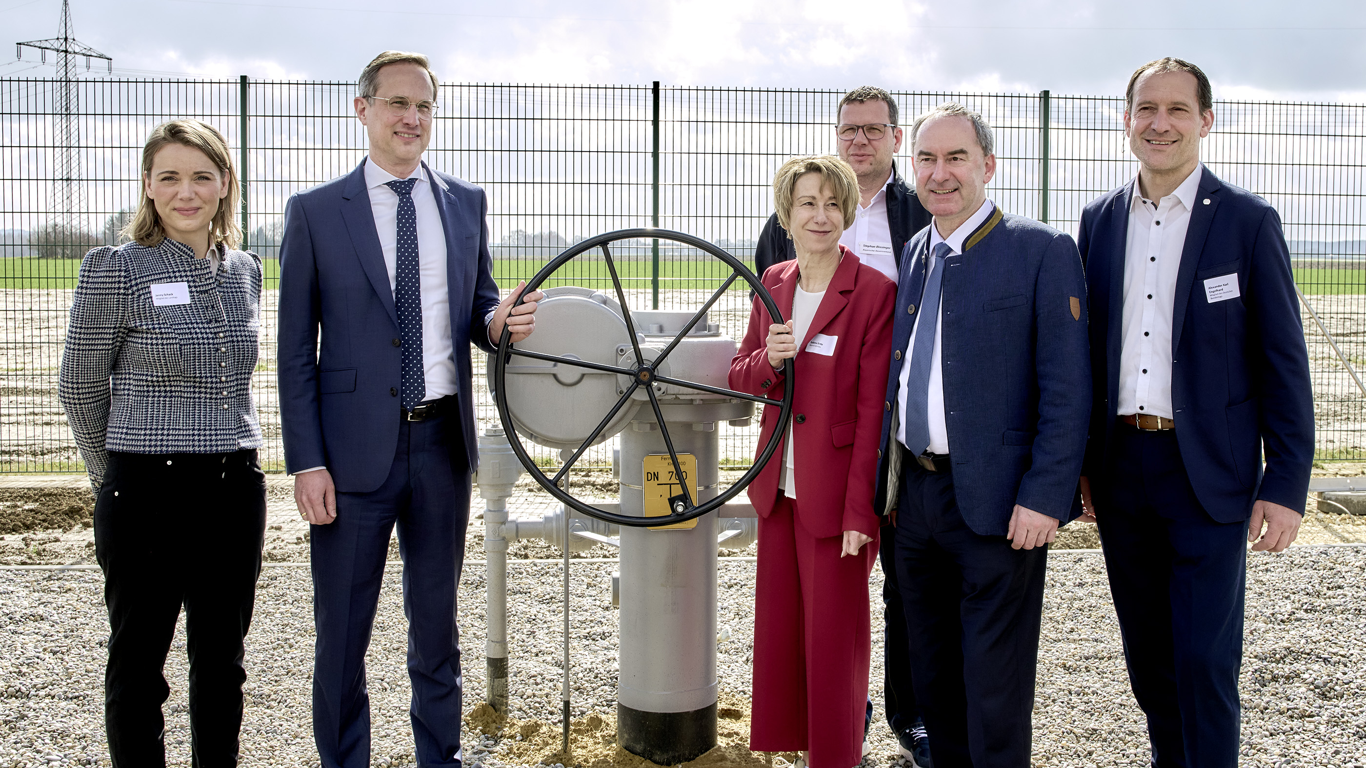 A group of people standing next to an industrial valve at an outdoor setting with a metal fence and sky in the background.