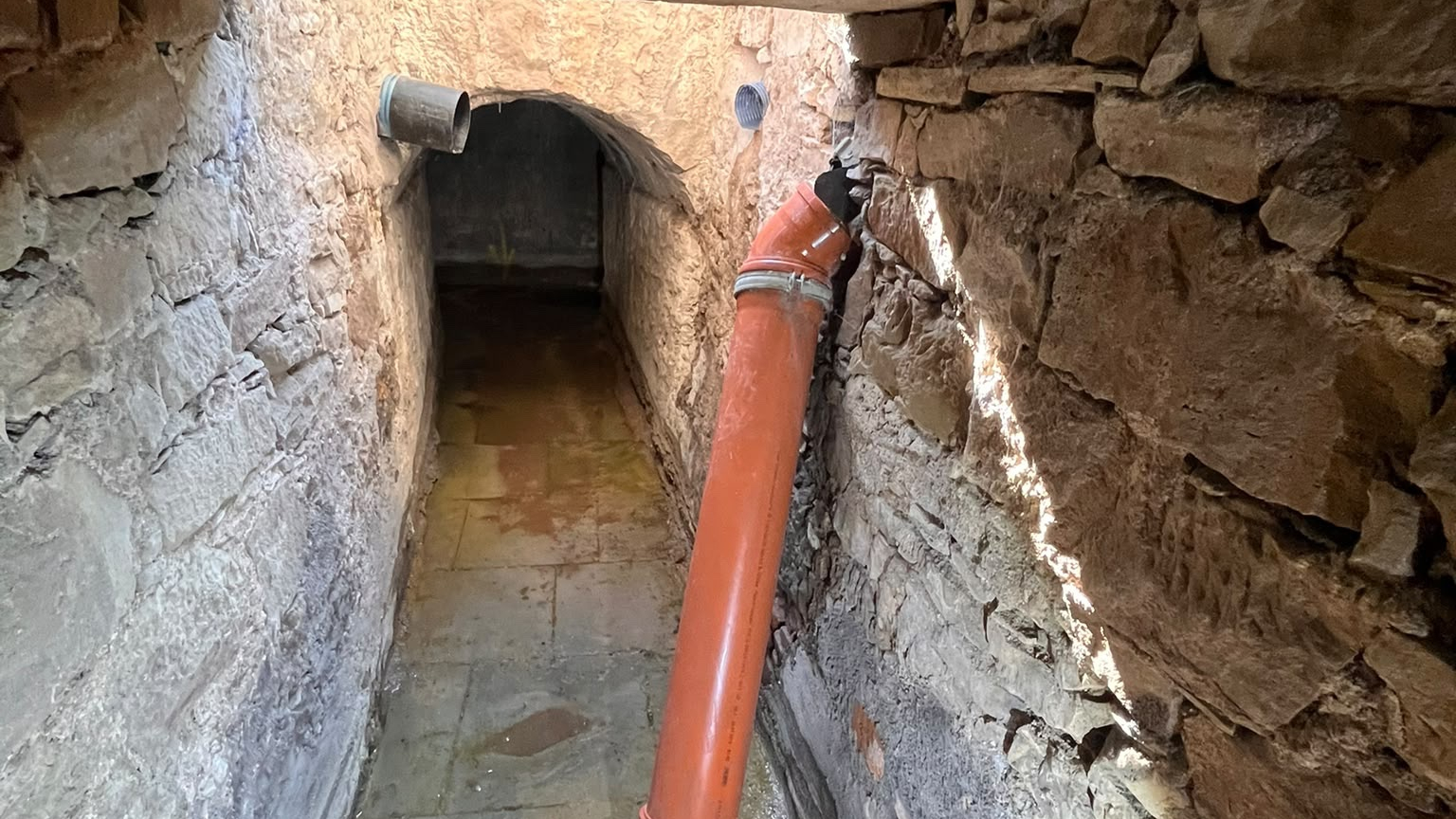Narrow stone drainage tunnel with shallow water on the floor and an orange pipe extending from the wall.
