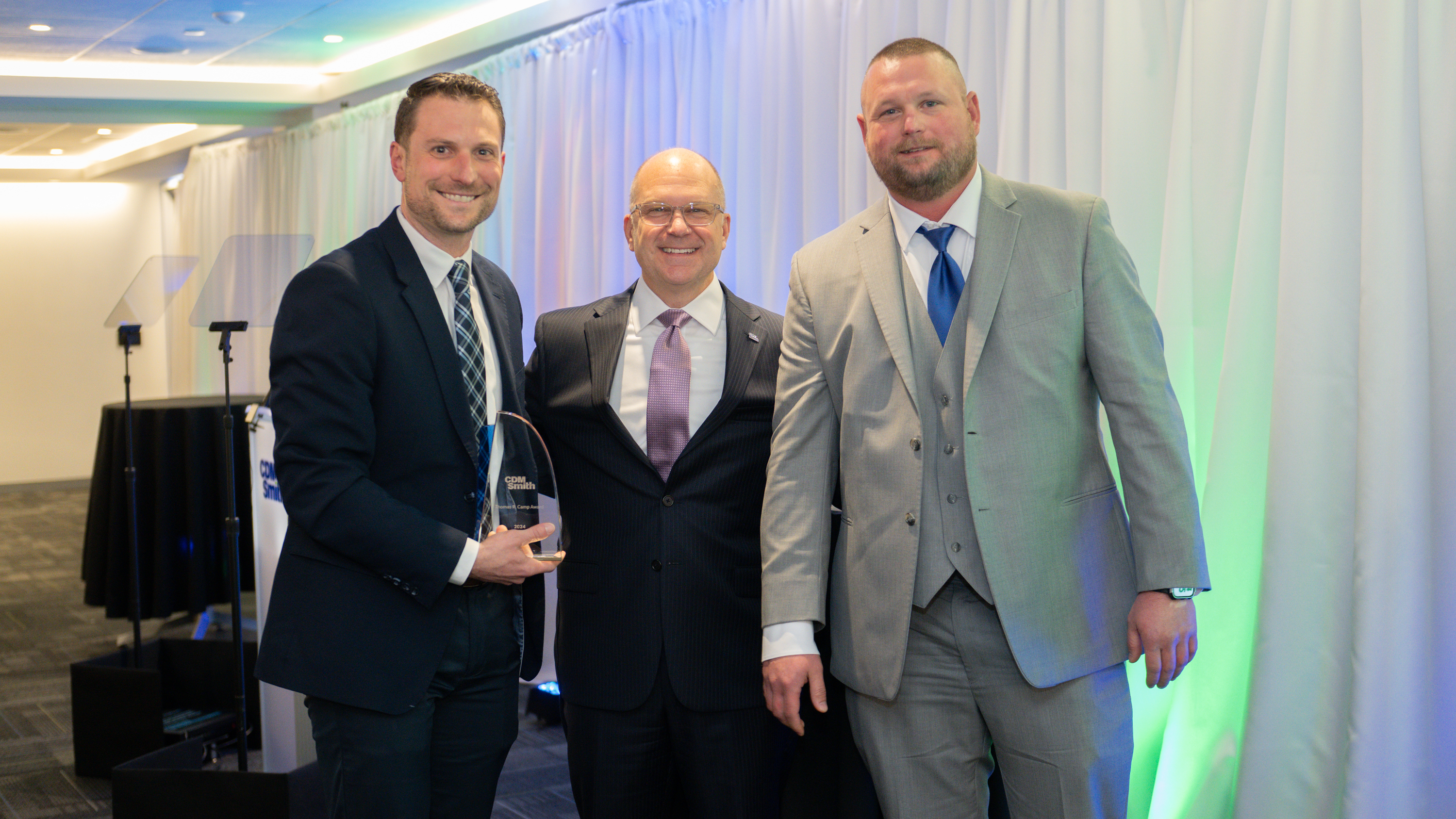 Three men in suits at an award ceremony; one holds a trophy in his right hand.