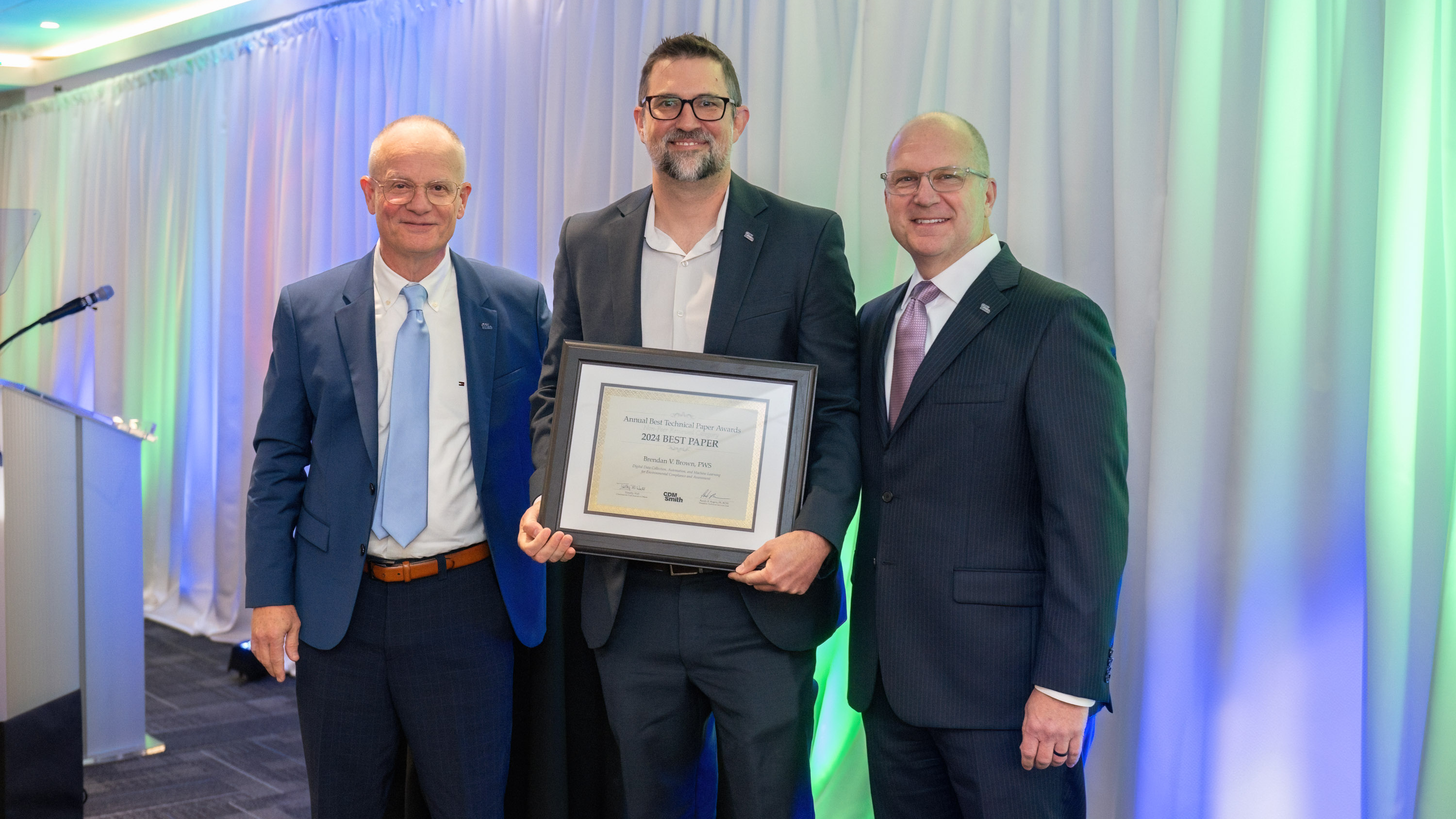 Three men in suits stand together, one holding a framed award titled "2021 Best Paper."