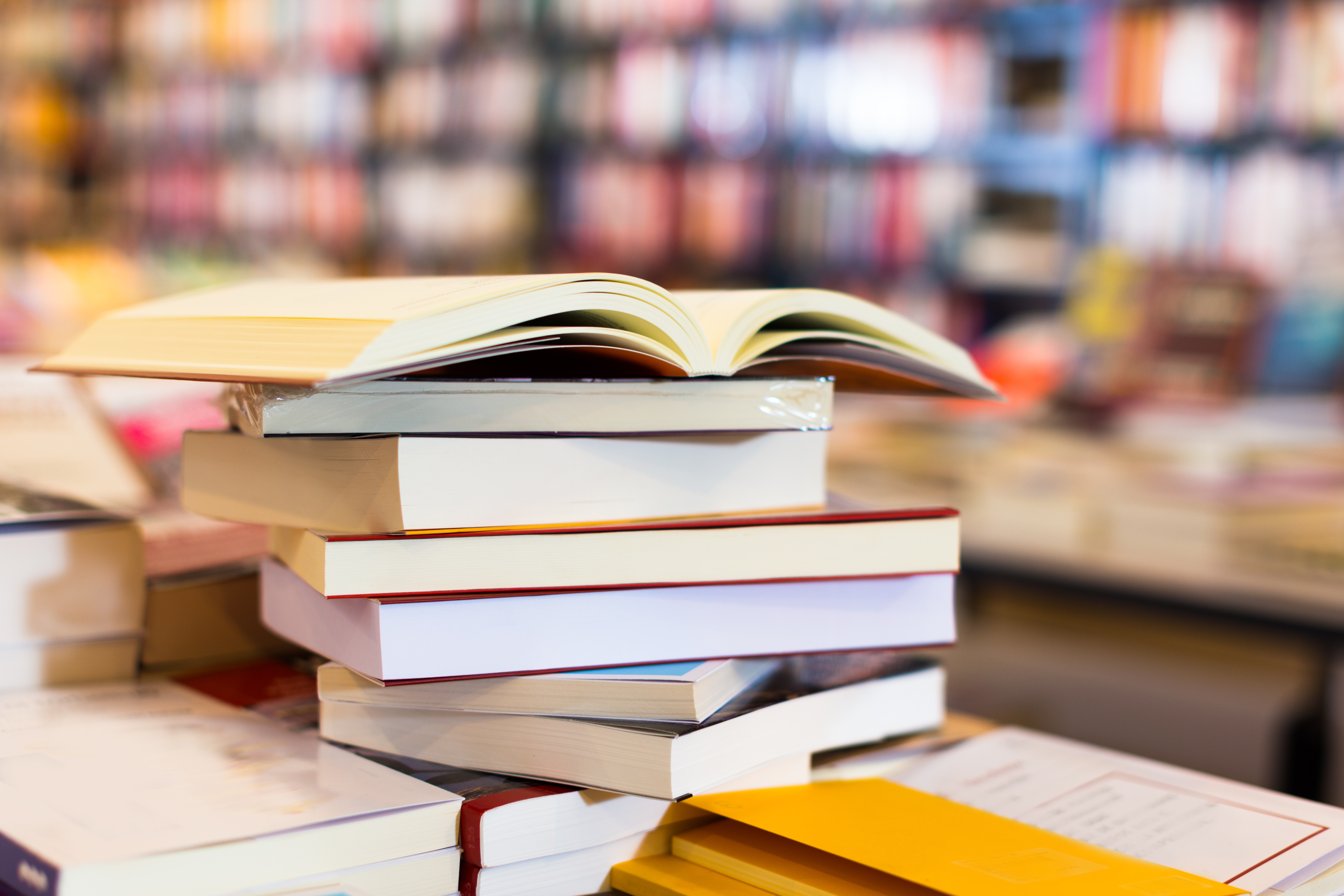 A stack of books with an open book on top, placed on a table in a bookstore or library with shelves in the background.
